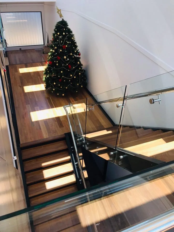 A decorated Christmas tree at the top of a staircase in a house, with sunlight casting shadows on the wooden floor.