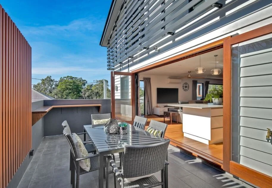 Outdoor balcony with a dining table and chairs, view into a modern living room with a flat-screen TV, kitchen island, and contemporary furnishings, during daytime with blue sky