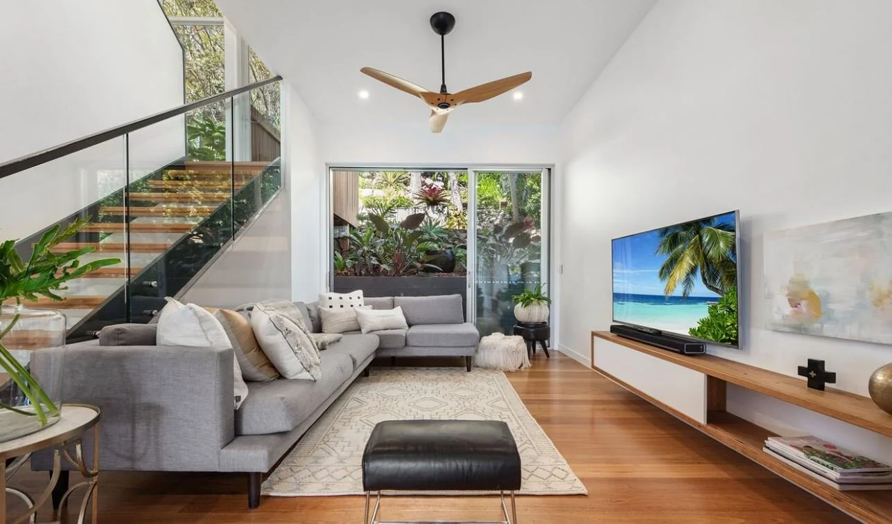 Modern living room with gray sectional sofa, glass staircase, wooden flooring, ceiling fan, tv showing a beach scene, and large window with greenery outside.