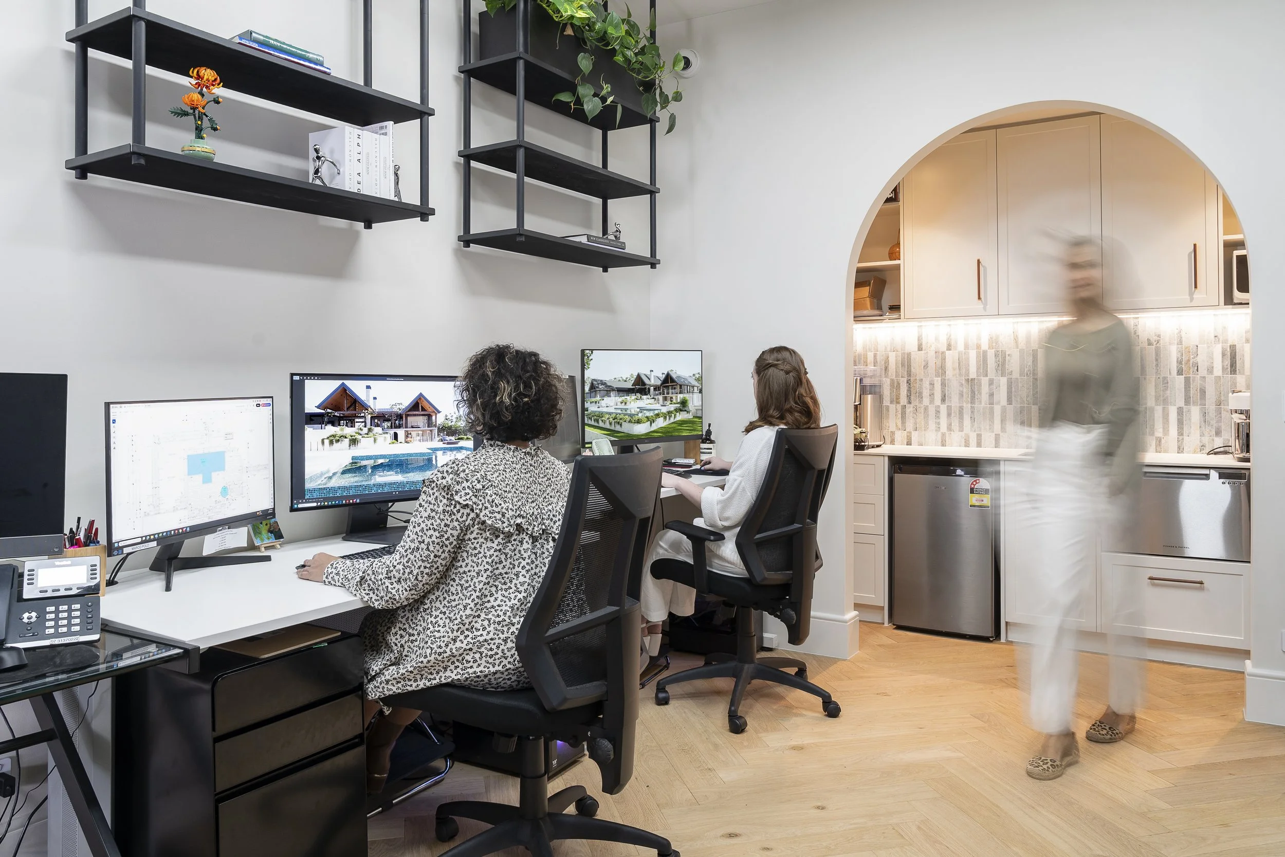 Two women working at desks with dual monitors in a home office, with kitchen visible through an arched doorway. A blurred person walks past in the foreground.