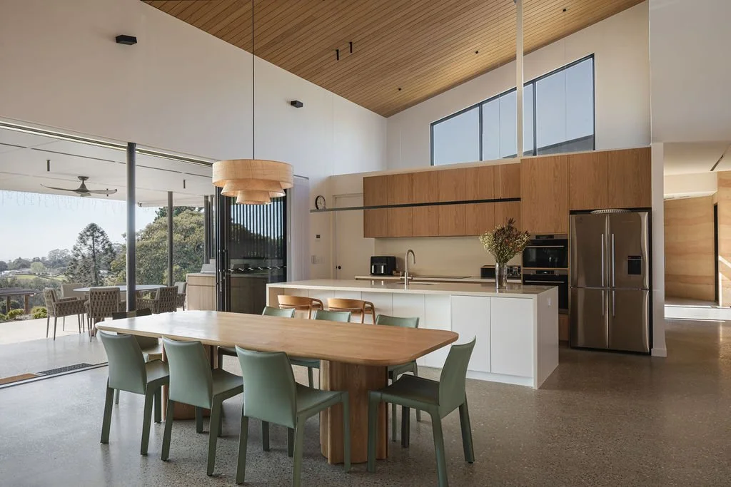 Modern kitchen with wooden cabinetry, white island, stainless steel refrigerator, dining table with green chairs, and large windows showing outdoor scenery.