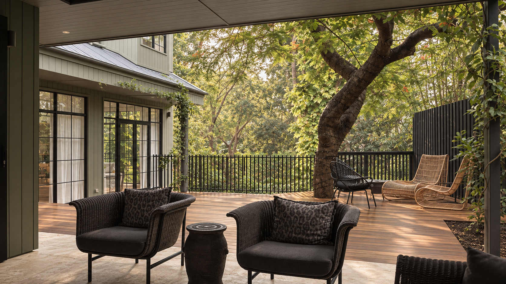 Outdoor patio with wicker chairs and leopard-print cushions, overlooking a lush garden with trees, a wooden deck, and modern house with large glass doors.