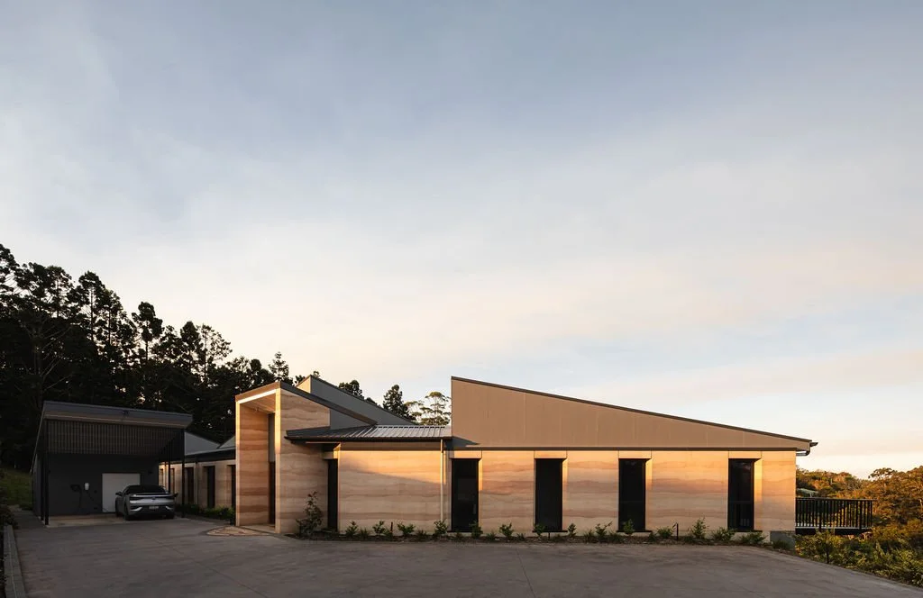 Modern single-story house with sloped roof, wooden exterior, large windows, and a garage with a car, set against a backdrop of trees and a clear sky at sunset.
