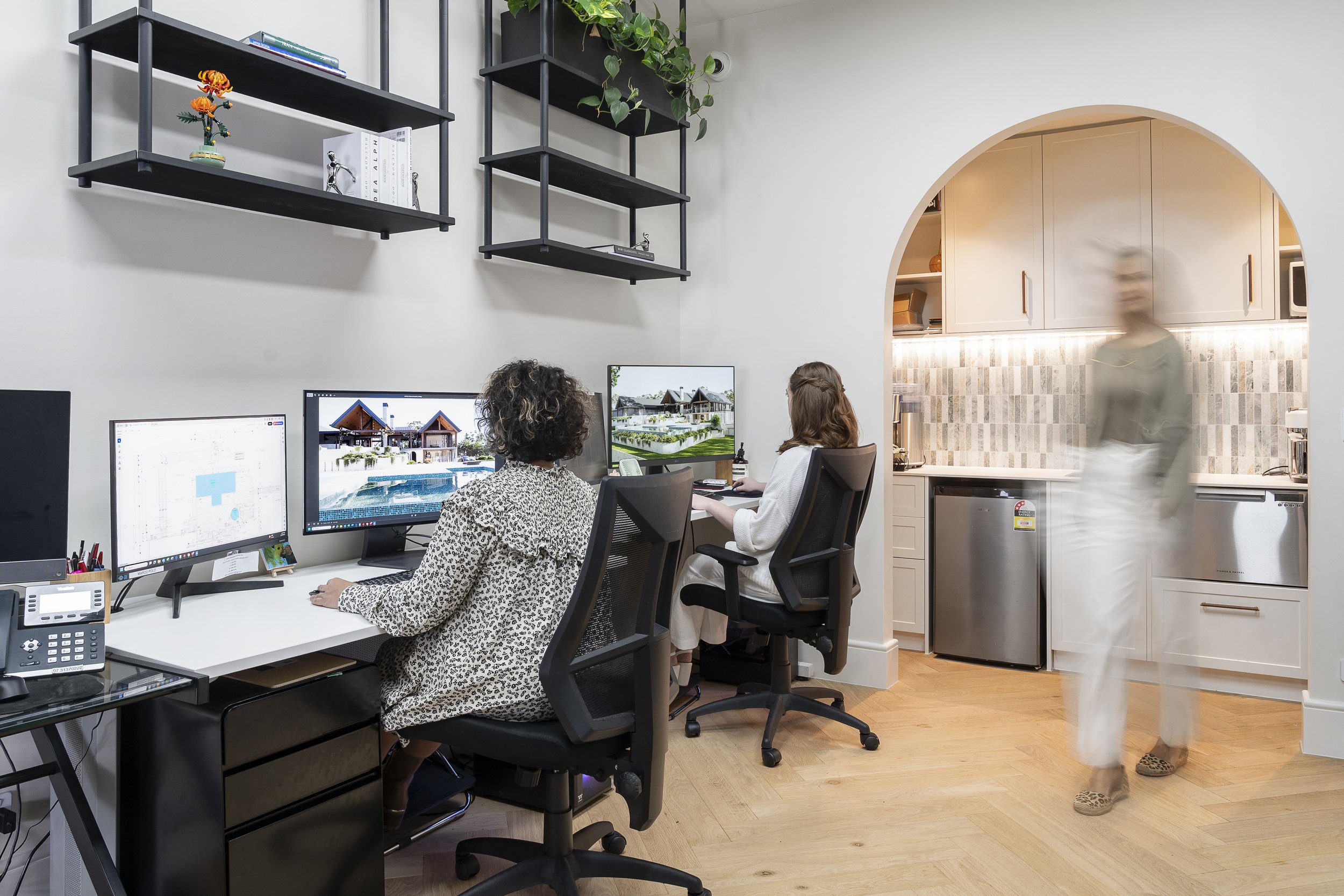 Two women working at desks with computer monitors in a modern home office. A woman with curly hair and a leopard print shirt is seen from behind, while a woman with straight hair and white clothing is working in front of a large monitor. In the background, there is a kitchen with beige cabinetry, an arched opening, and a blurred figure walking by, wearing leopard print slippers.