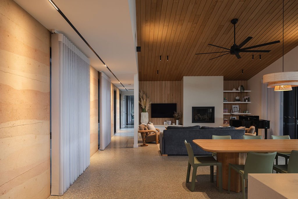 Modern living room with wooden ceiling, black ceiling fan, fireplace, TV, bookshelf, concrete floor, large dining table, and upholstered chairs.