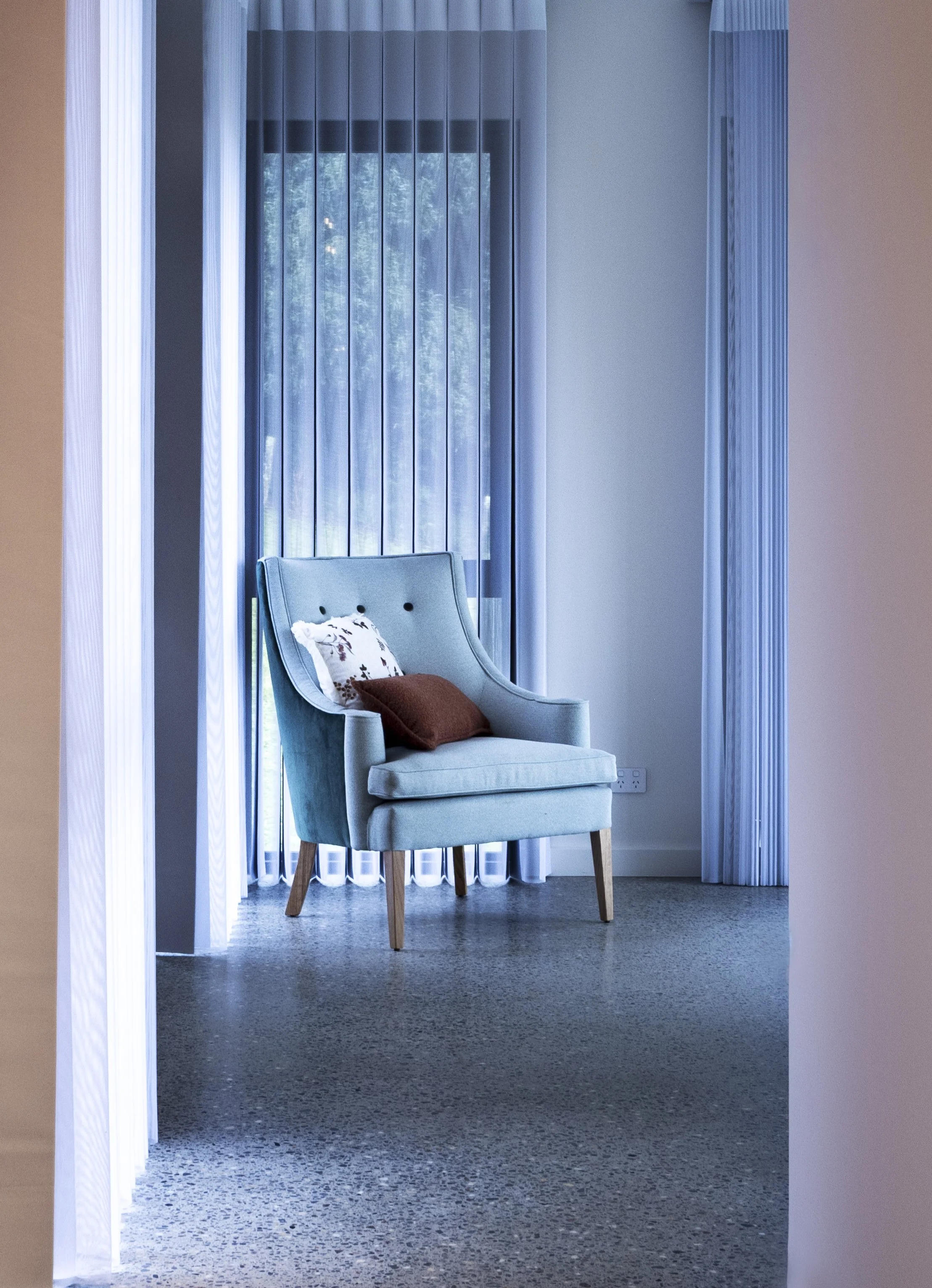 A light blue armchair with a floral pillow and brown pillow, positioned in front of a large window with vertical blinds, in a room with beige walls and gray carpet.