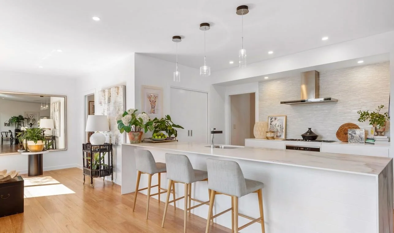 Modern kitchen with white cabinets, a long island with three gray barstools, decorative vases, and plants, connected to a dining area with a large mirror and wooden floors.
