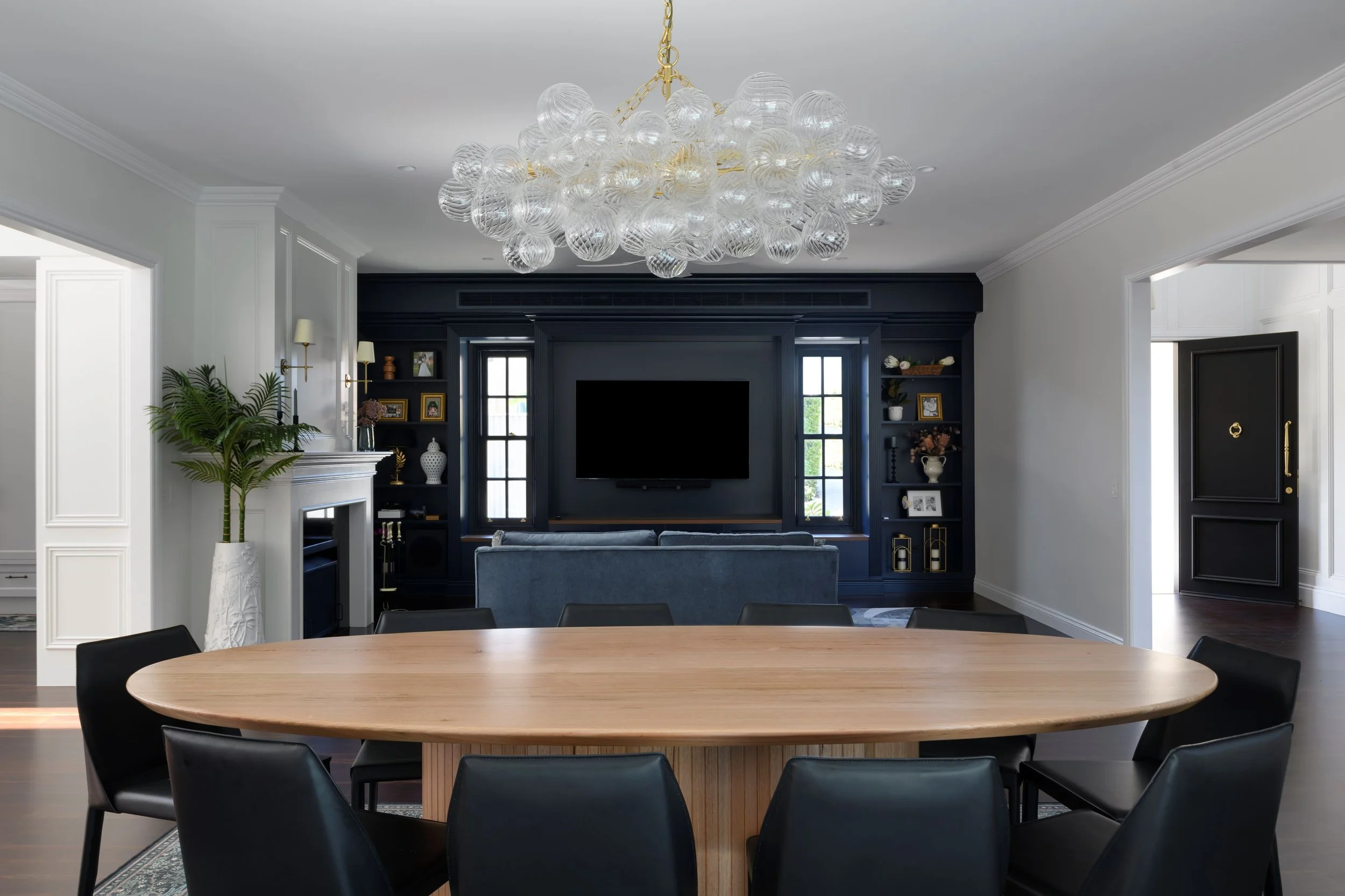 Modern living room with a round wooden dining table and black chairs, a chandelier with glass globes, a navy blue entertainment center with a TV, shelves with framed photos and decor, windows letting in natural light, and a fireplace with a potted pl