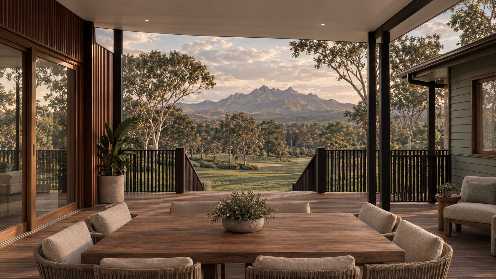 View from a covered porch with wooden flooring, railing, and furniture overlooking a lush green landscape with trees and mountains in the distance during sunset.