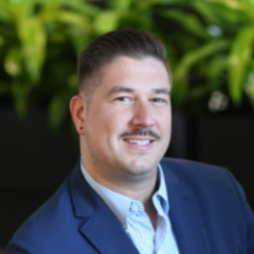 Portrait of a man in a blue suit and light blue shirt, smiling, with green plants in the background.