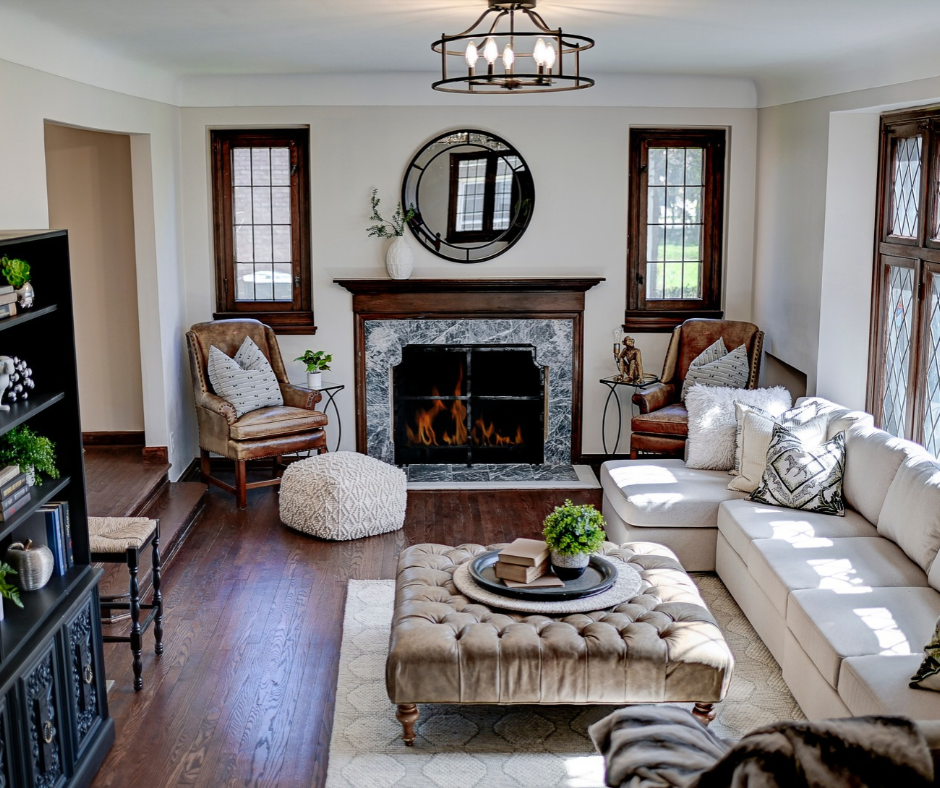 Cozy, well-lit living room featuring hardwood floors, a stone fireplace, leather accent chair, tufted ottoman, and traditional wooden window frames.