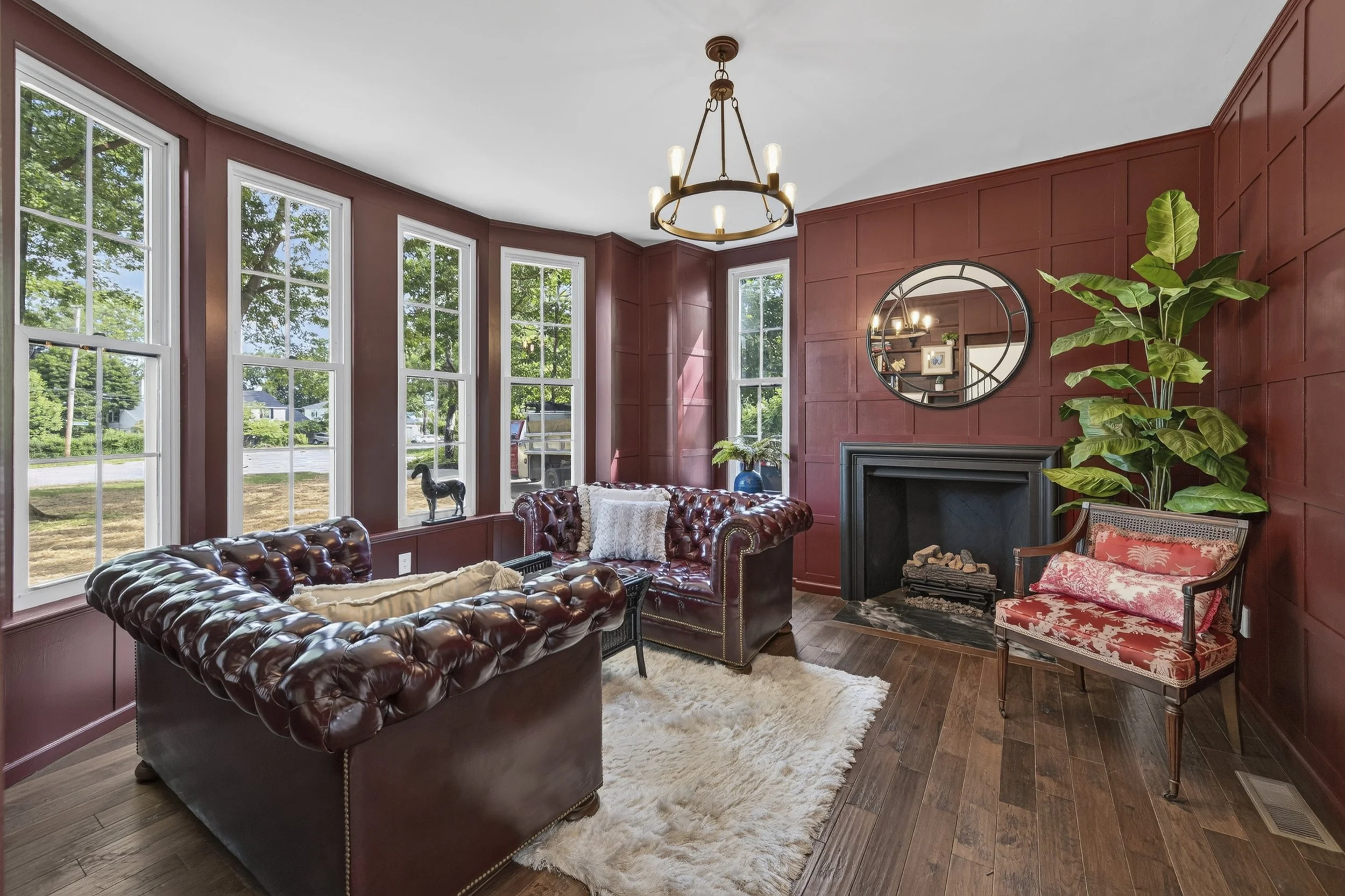 Moody burgundy sitting room with board and batten paneling, leather Chesterfield sofas, black fireplace, and bay windows by VNTG Home