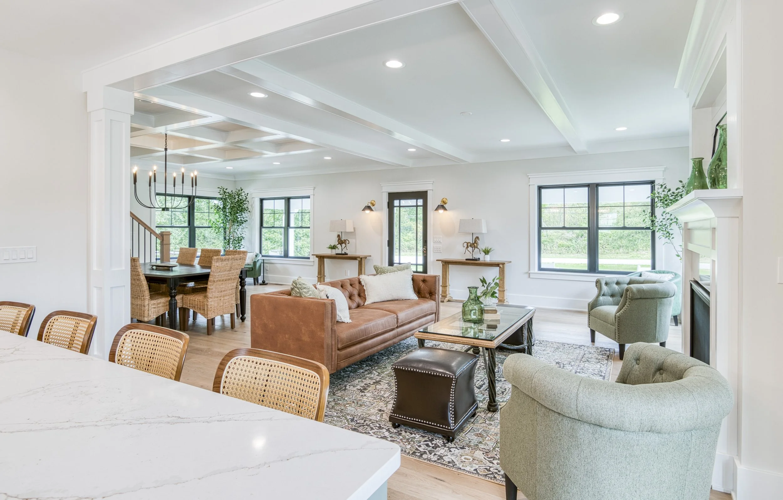 Bright living room with beige sofa, armchairs, wooden console tables with lamps, large windows, and greenery outside, featuring a cozy and modern decor.