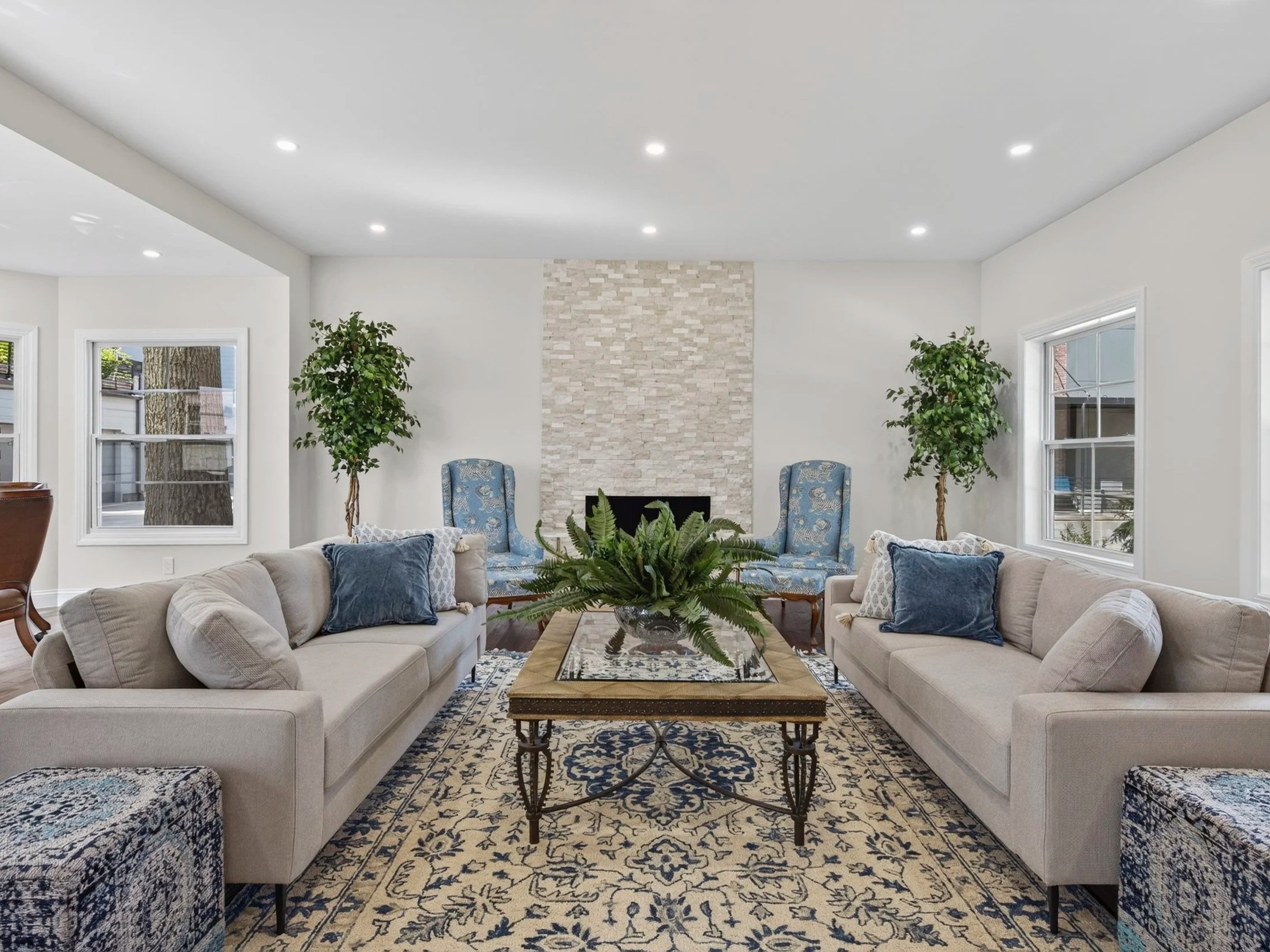 A spacious living room with two off-white sofas facing each other, a rectangular glass-top coffee table with a decorative metal base in the center, blue patterned accent chairs, a brick fireplace, two large potted trees, and large windows allowing natural light.