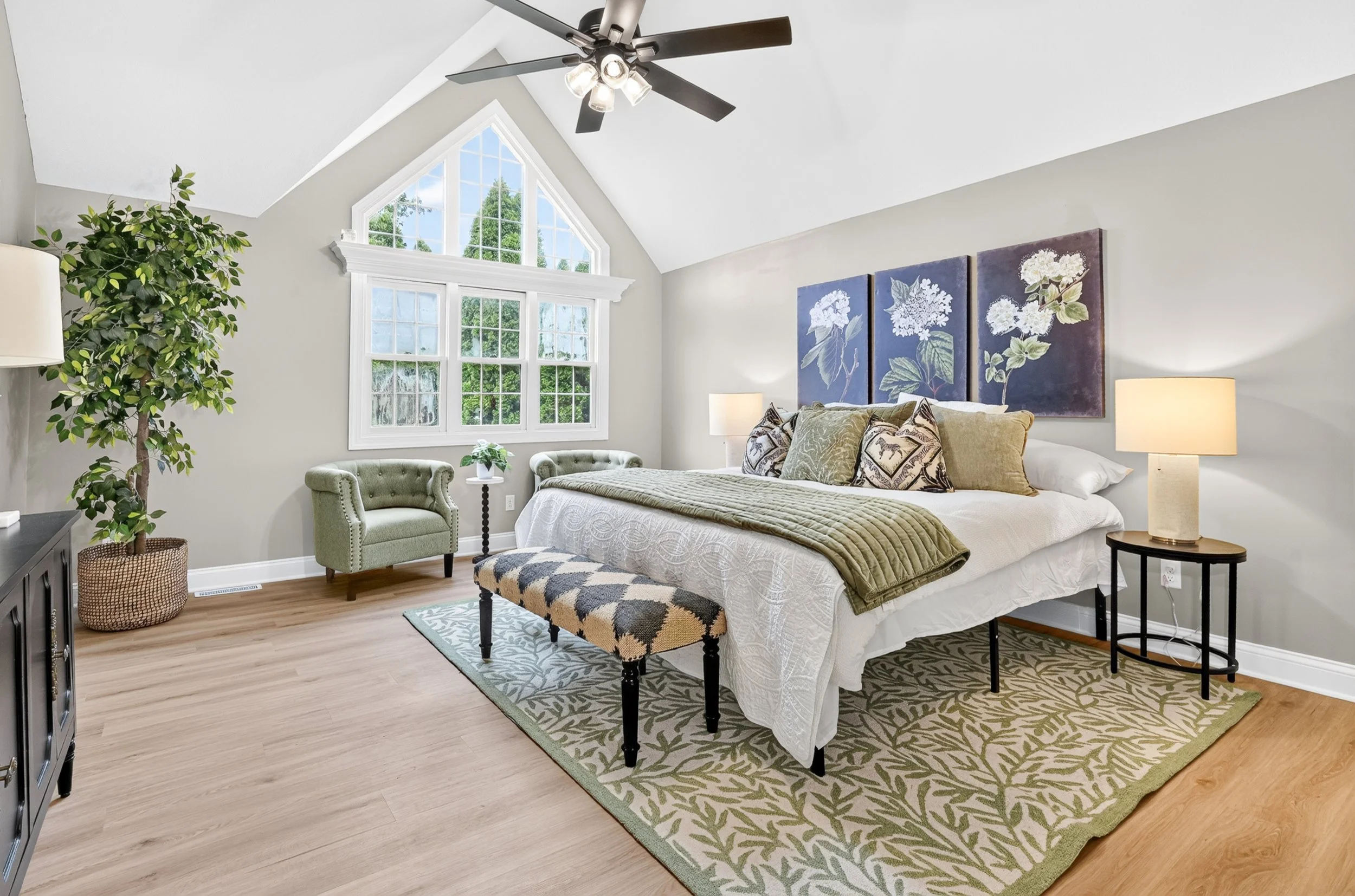 Bright bedroom with a large triangular window, a bed with decorative pillows, two nightstands with lamps, a painting of white flowers on the wall, a green rug with leaf patterns, and a tall indoor plant.