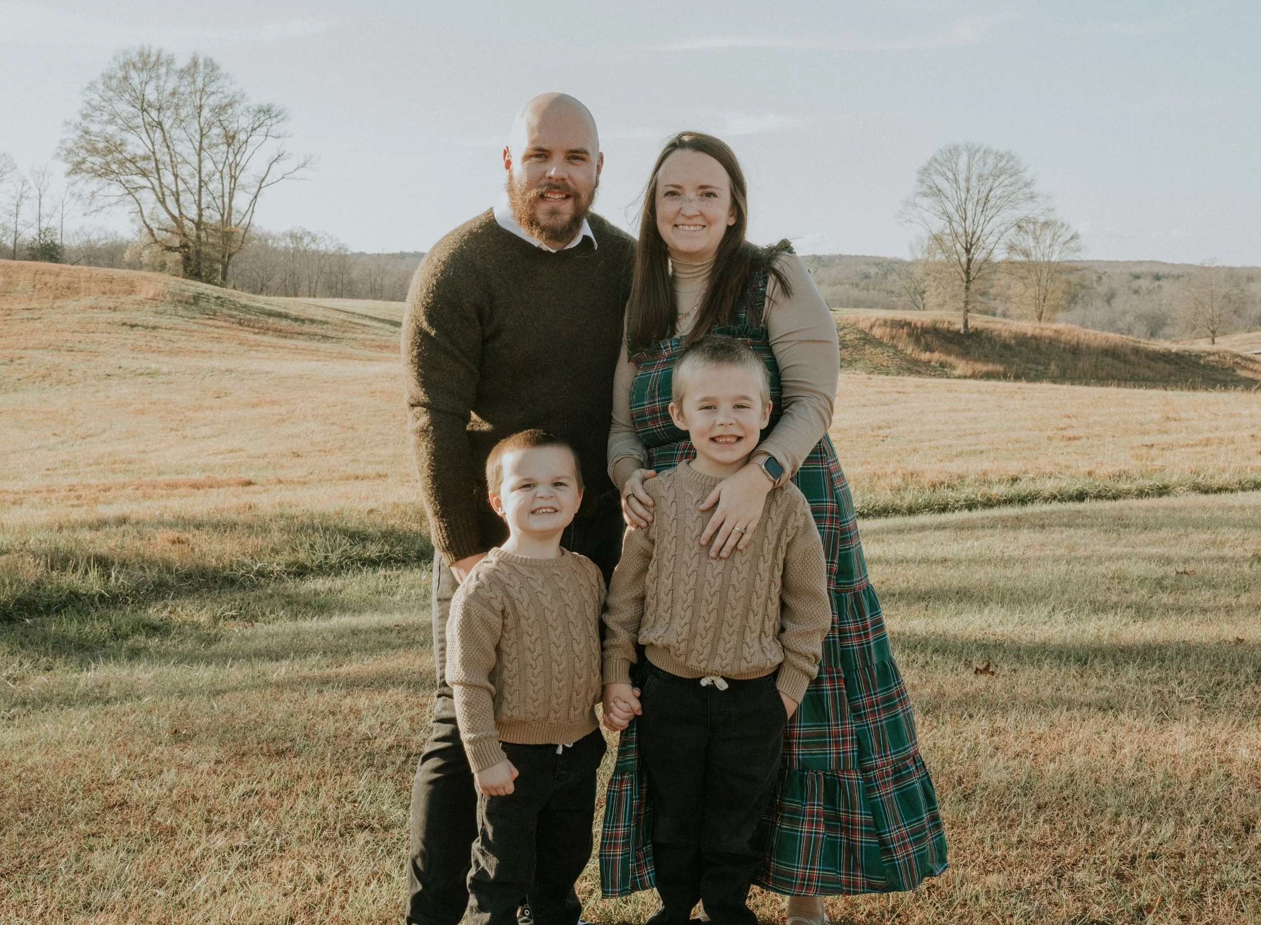 A family of five standing outdoors in a grassy field with trees in the background, smiling at the camera during daytime.