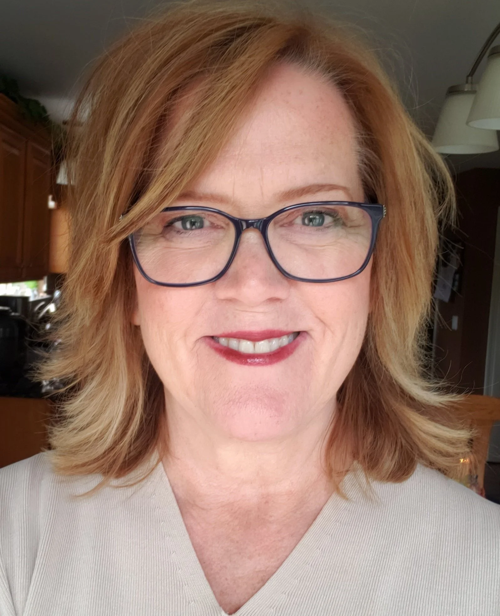 Close-up photo of a woman with red hair and glasses, smiling indoors.