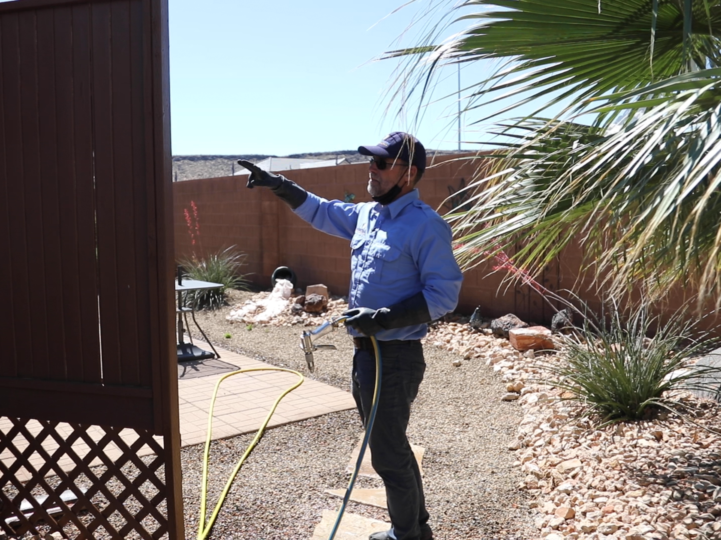 A man wearing sunglasses, gloves, and a blue long-sleeve shirt is using a hose to spray or wash a backyard with desert landscaping, including rocks, plants, and a wooden fence.
