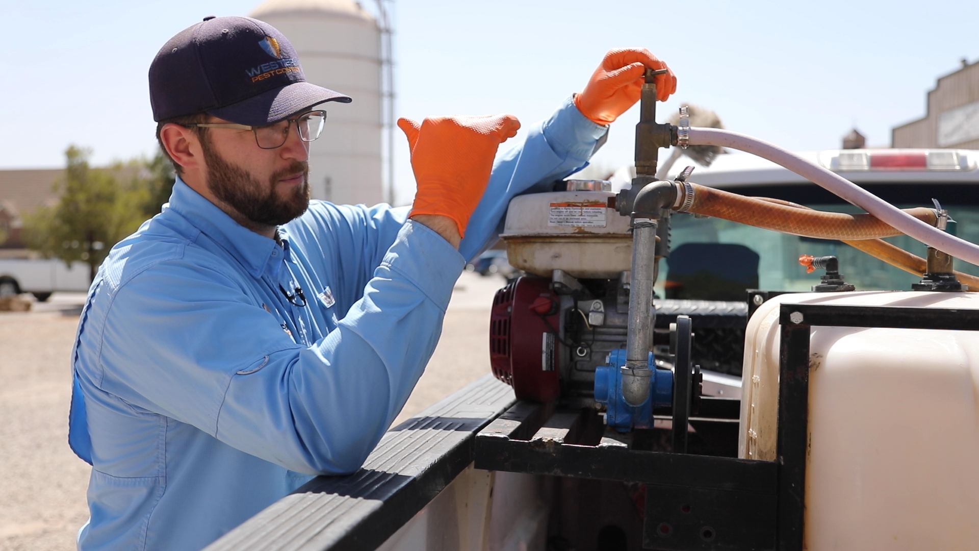 A man wearing an orange glove, glasses, a navy cap, and a blue shirt working on a piece of equipment outdoors, with trees and buildings in the background.