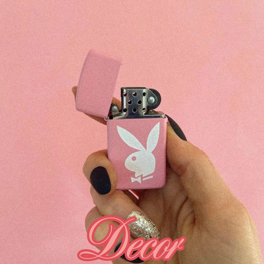 A pink lighter with a white Playboy bunny logo, held by a person with black nail polish and a gold glittery nail, against a pink background. The word 'Decar' is written in cursive at the bottom.