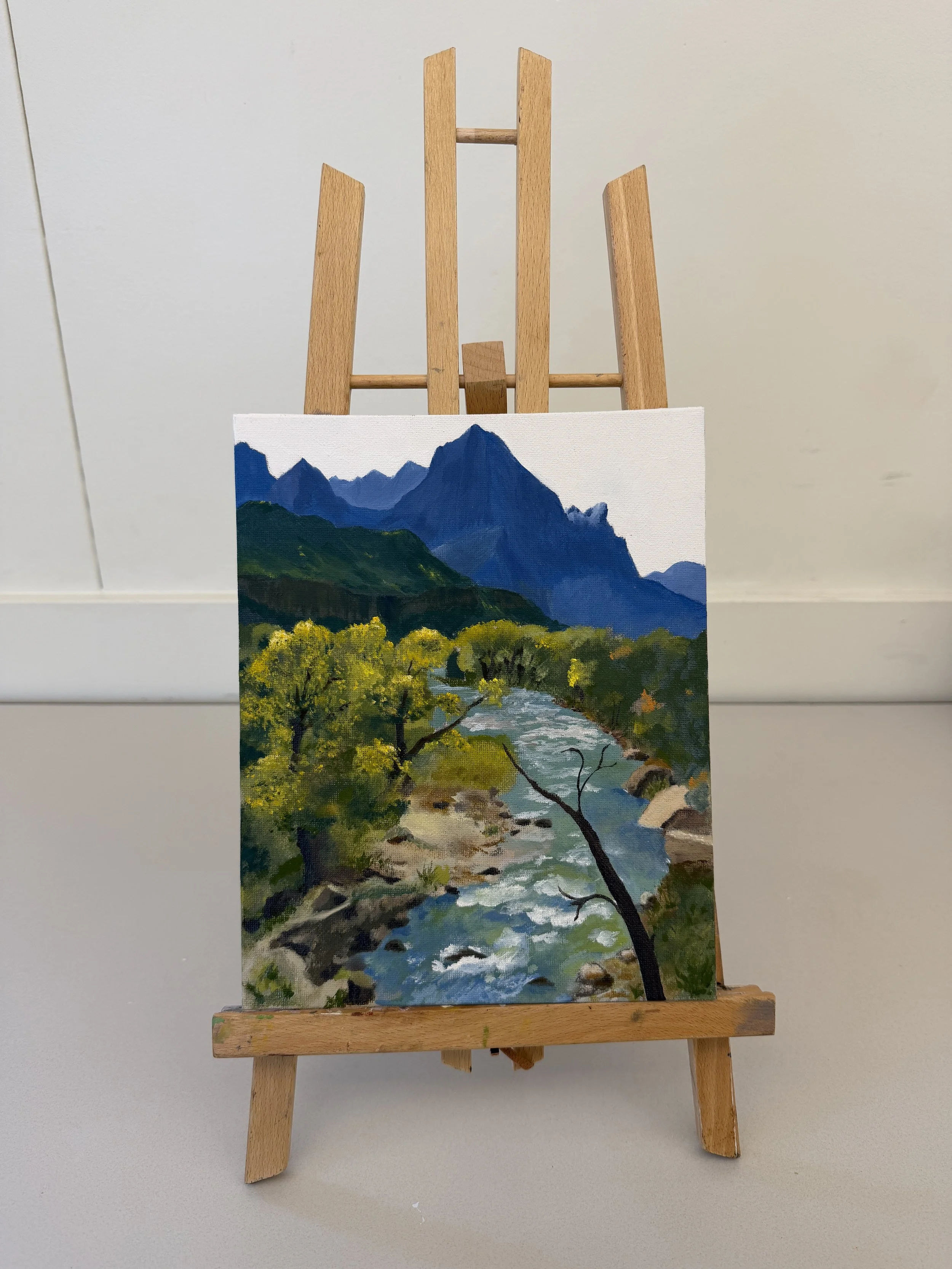 Painting of a mountain landscape with a river and trees on an easel.