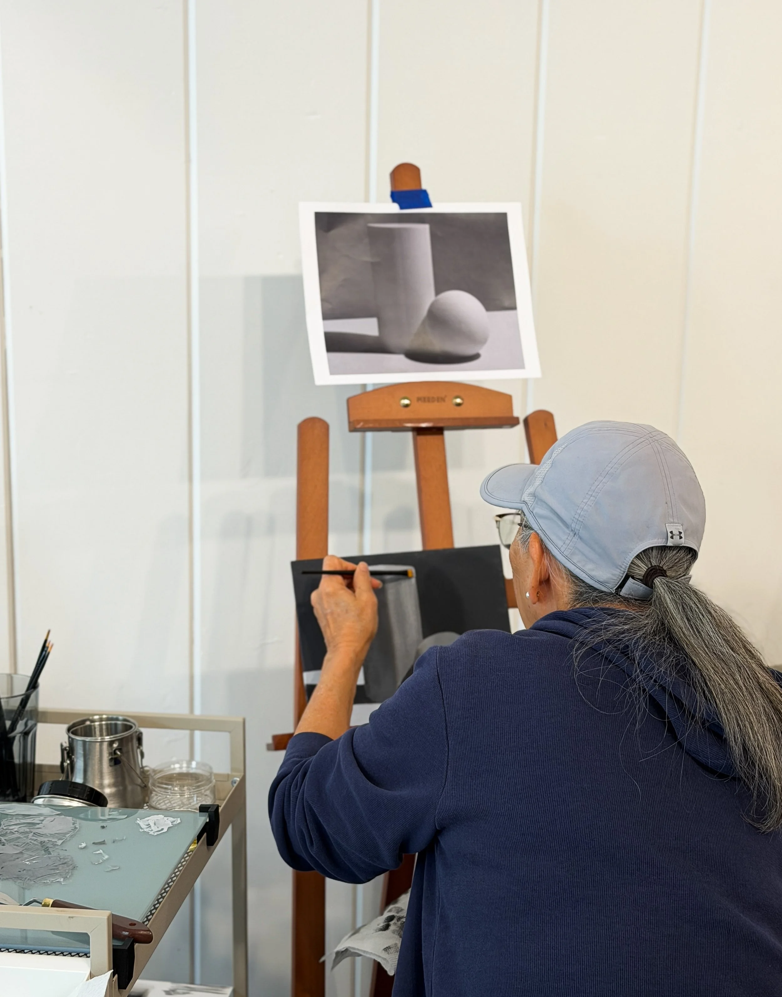 An artist or sculptor working on a sculpture in a studio, with a photograph of a white ceramic vessel and sphere on an easel in the background. The person is wearing a gray cap and navy blue hoodie and is seated at a worktable with sculpting tools and materials.