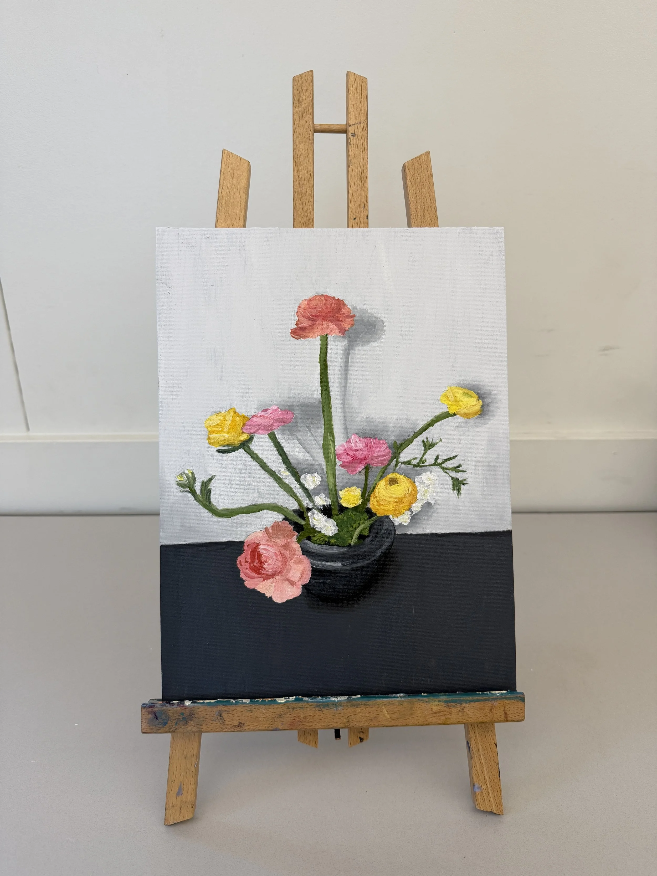 A still life painting of a bouquet of pink, yellow, and white flowers in a dark vase, set against a light gray background, displayed on an easel.