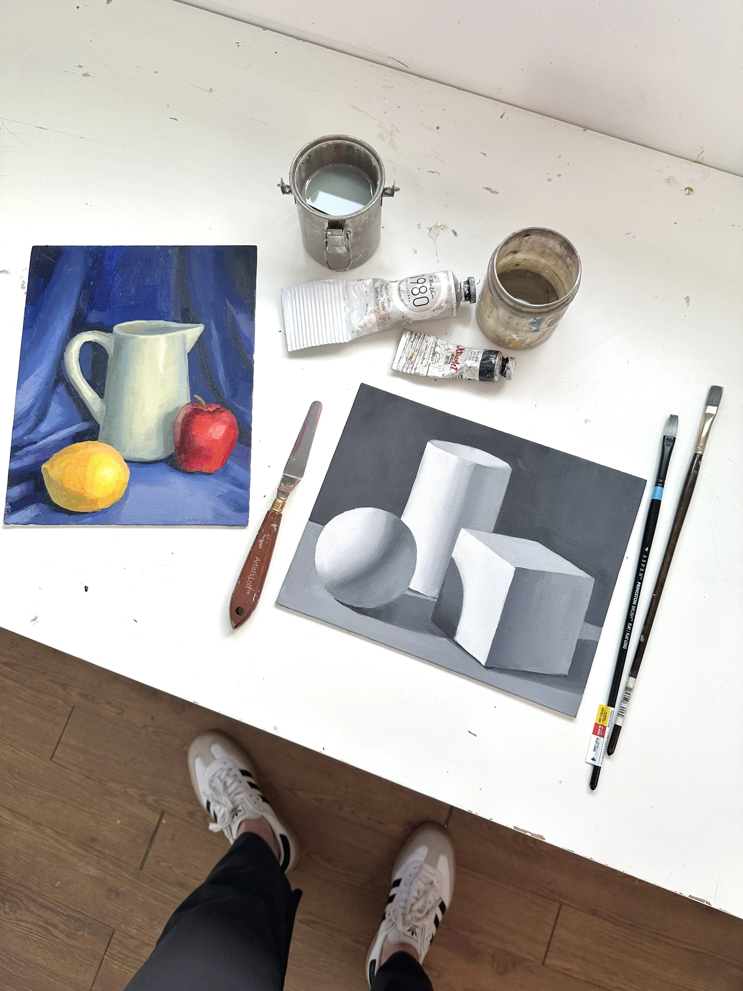 An artist's workspace featuring a painted still life with a pitcher, an apple, and a lemon on a mat, along with tubes of paint, paintbrushes, and a grayscale geometric painting on the table. There are also person's legs and feet in sneakers visible a