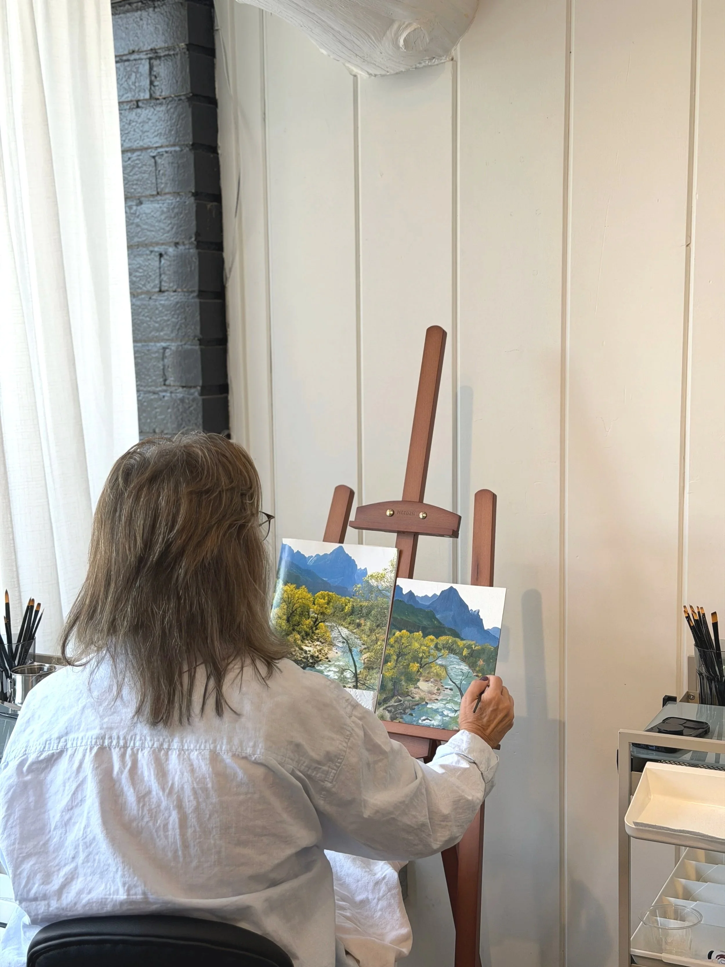 A woman with gray hair and glasses, wearing a white shirt, paints a landscape on a canvas with mountains, trees, and a river in a room with white walls and a black brick wall.