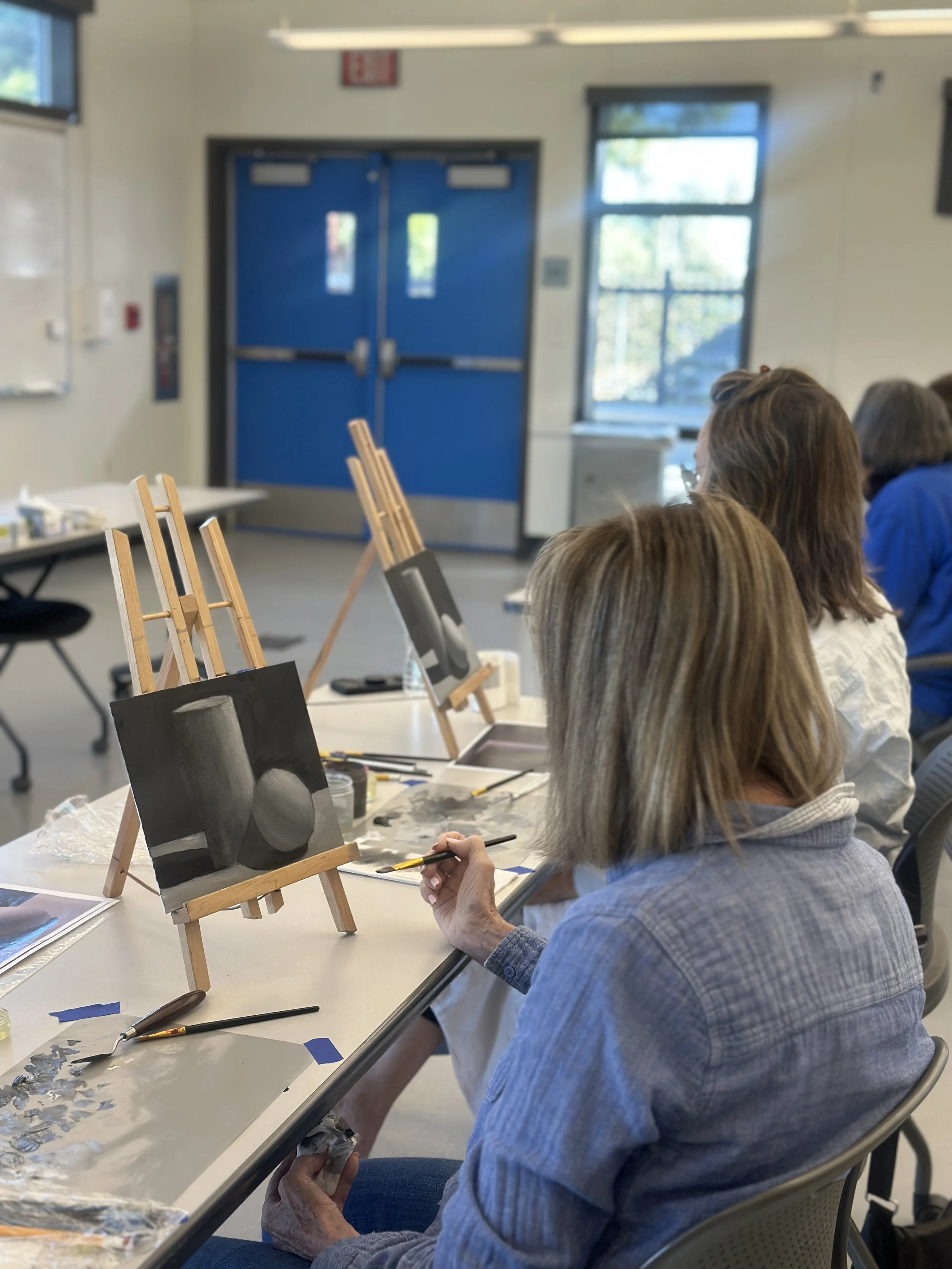 People participating in a painting class, working on small canvas paintings of a black cat. The room has large windows, blue doors, and art supplies on the table.
