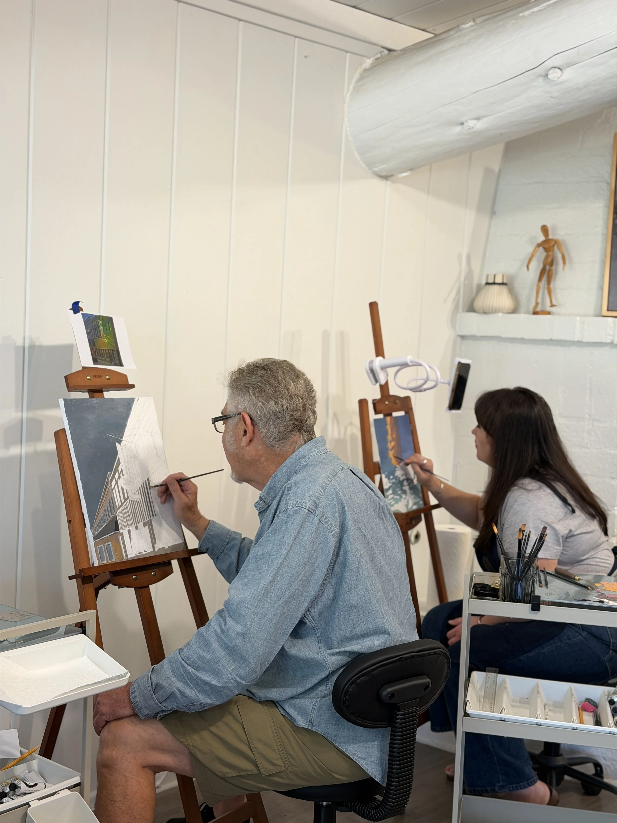 Two people painting canvases on easels in a bright apartment with white walls, one man and one woman, with art supplies nearby.