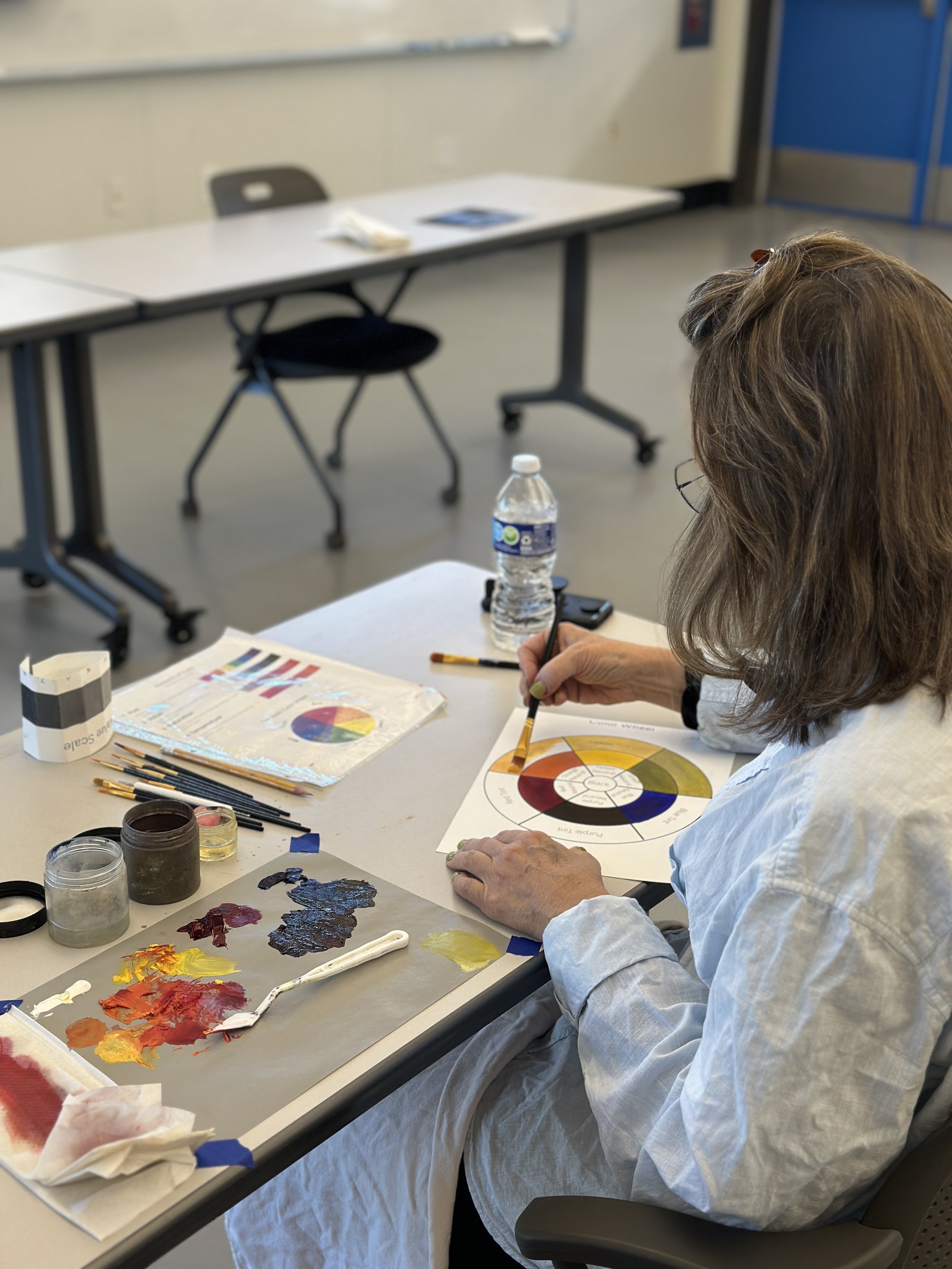 Person working on a color wheel painting project at a desk, surrounded by painting supplies and color swatches in a classroom or studio setting.
