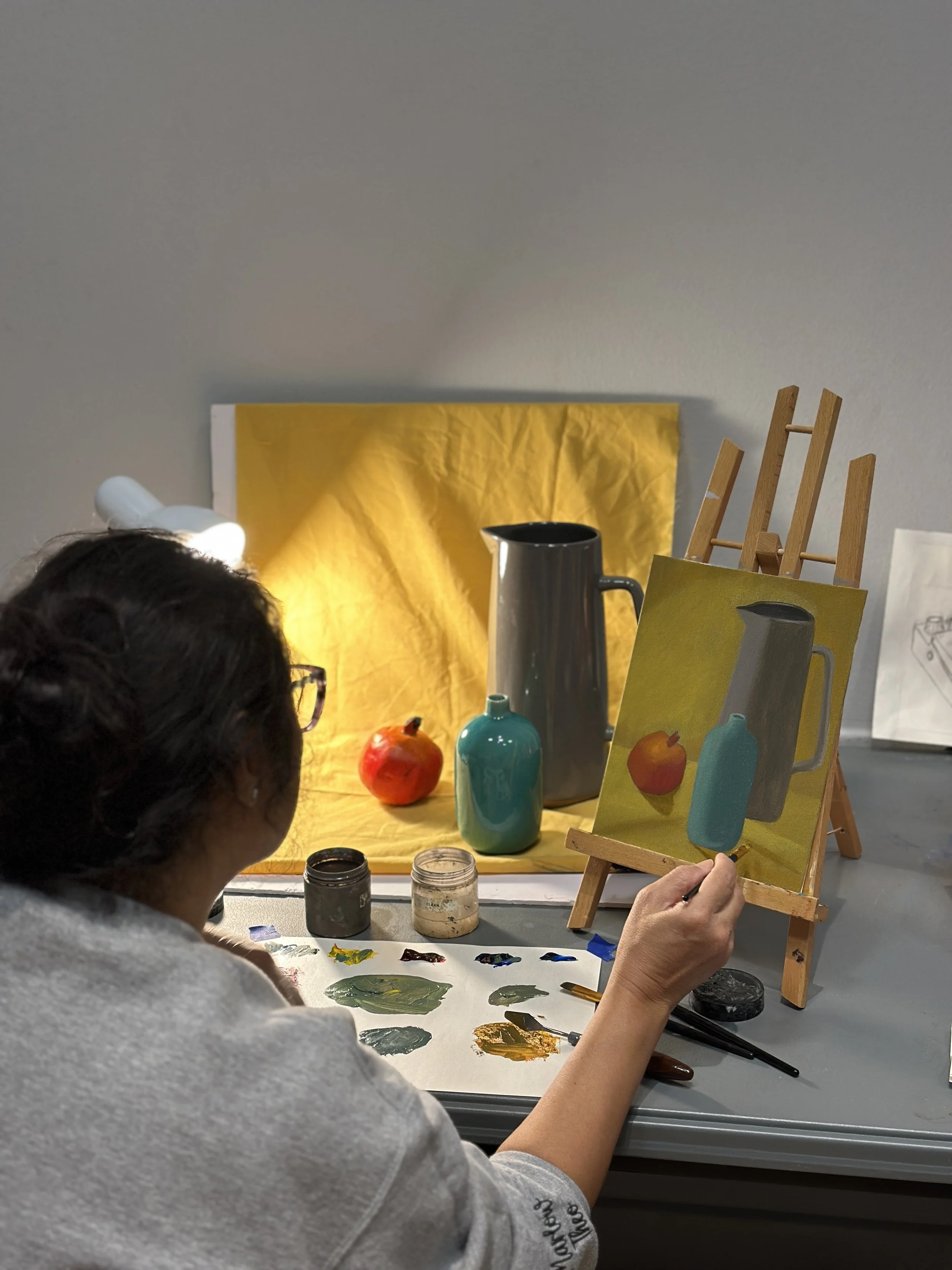 A person painting a still life on a small canvas set on an easel, with objects like a pomegranate and a ceramic jug arranged on a table, in a brightly lit art studio.