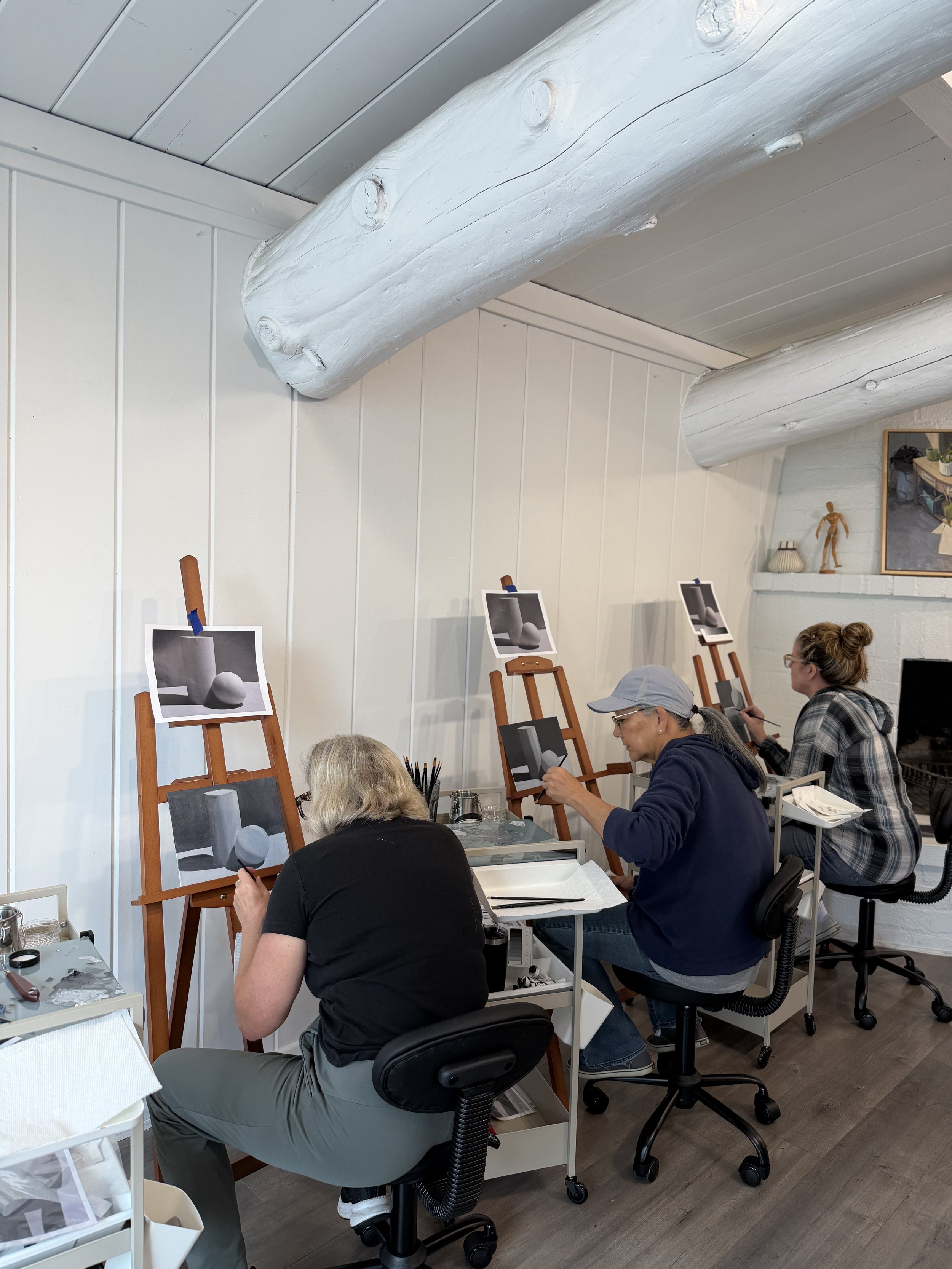 People sitting at tables in an art studio, painting sculptures on easels, with a white painted log and photographs of sculptures on the wall.