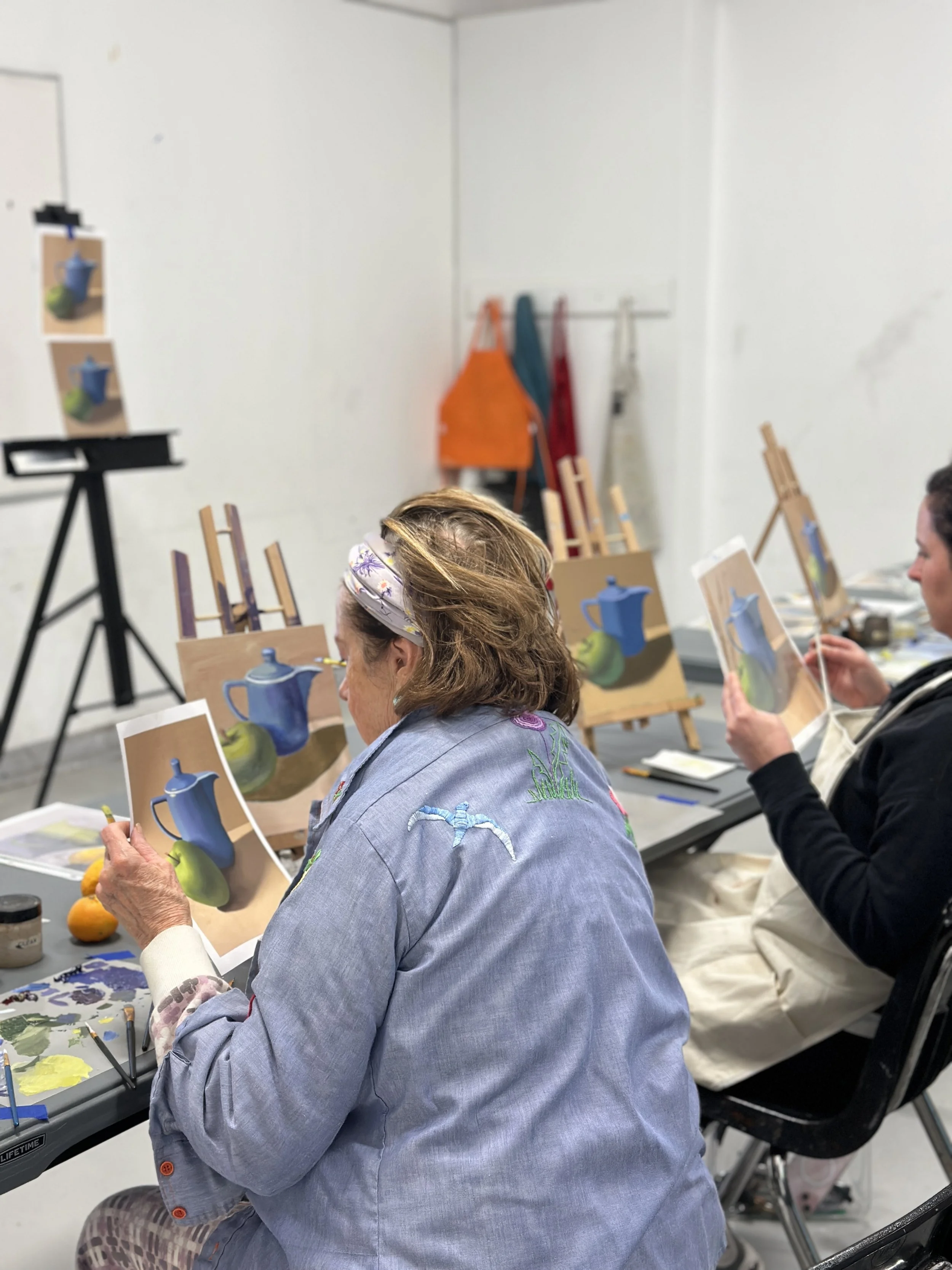 People painting still life pictures of teapots on canvases in an art class.