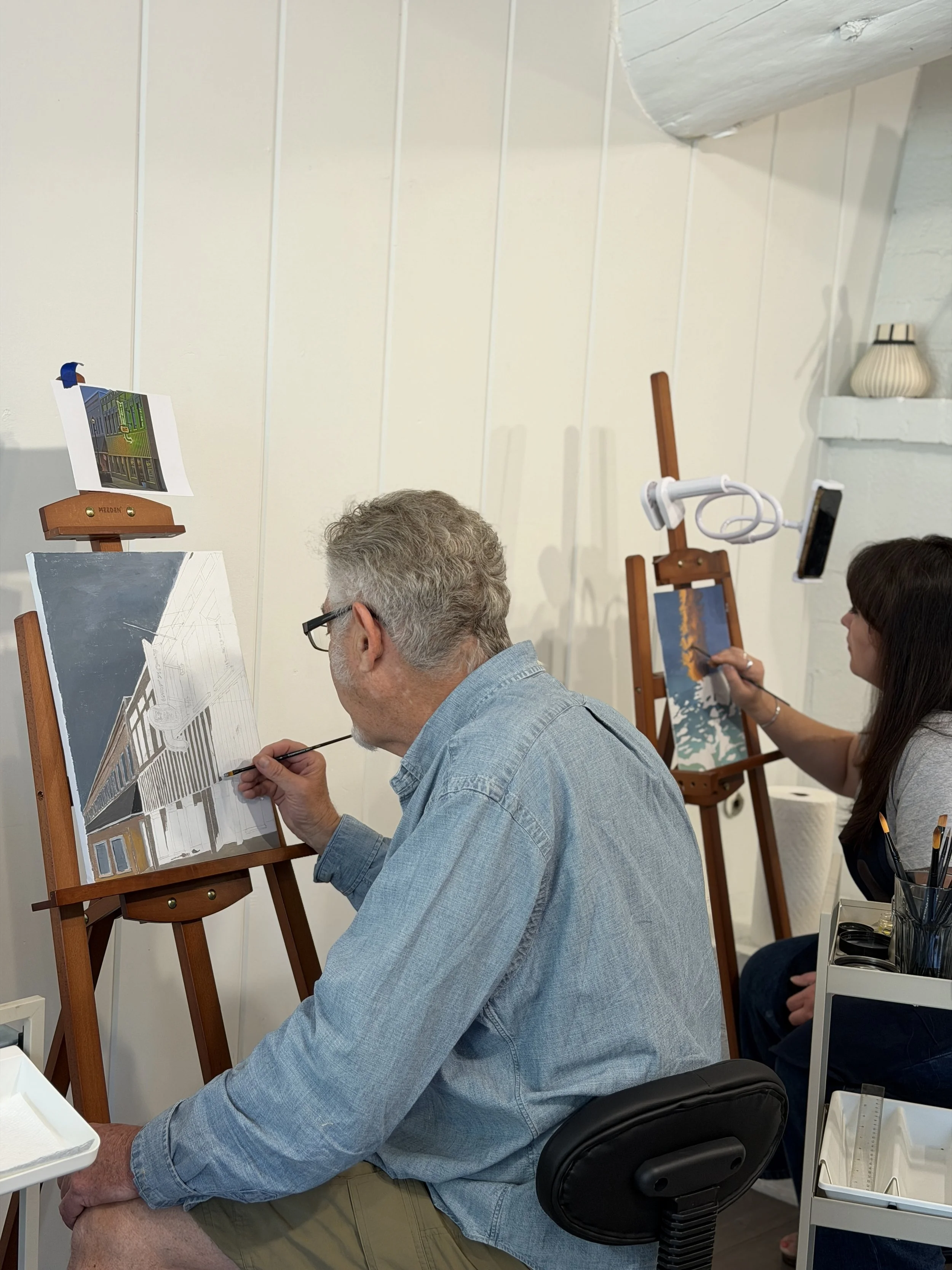 Two people painting house facades on easels in an art workshop, with art supplies and a white wall in the background.