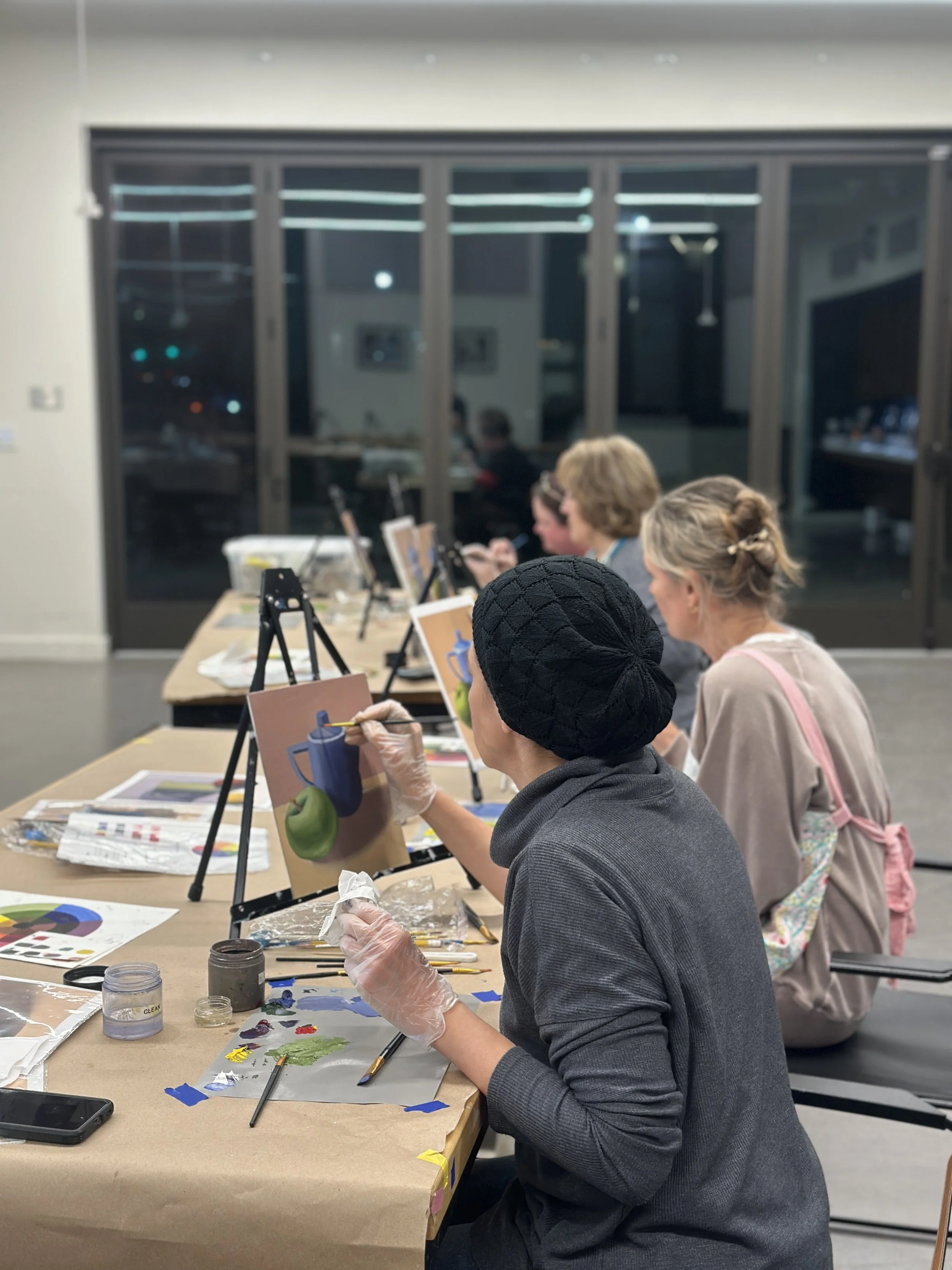 People engaged in a painting class, seated at a long table with art supplies such as paint, brushes, and palettes, focusing on their artwork. The background shows a large window and a room with various equipment.