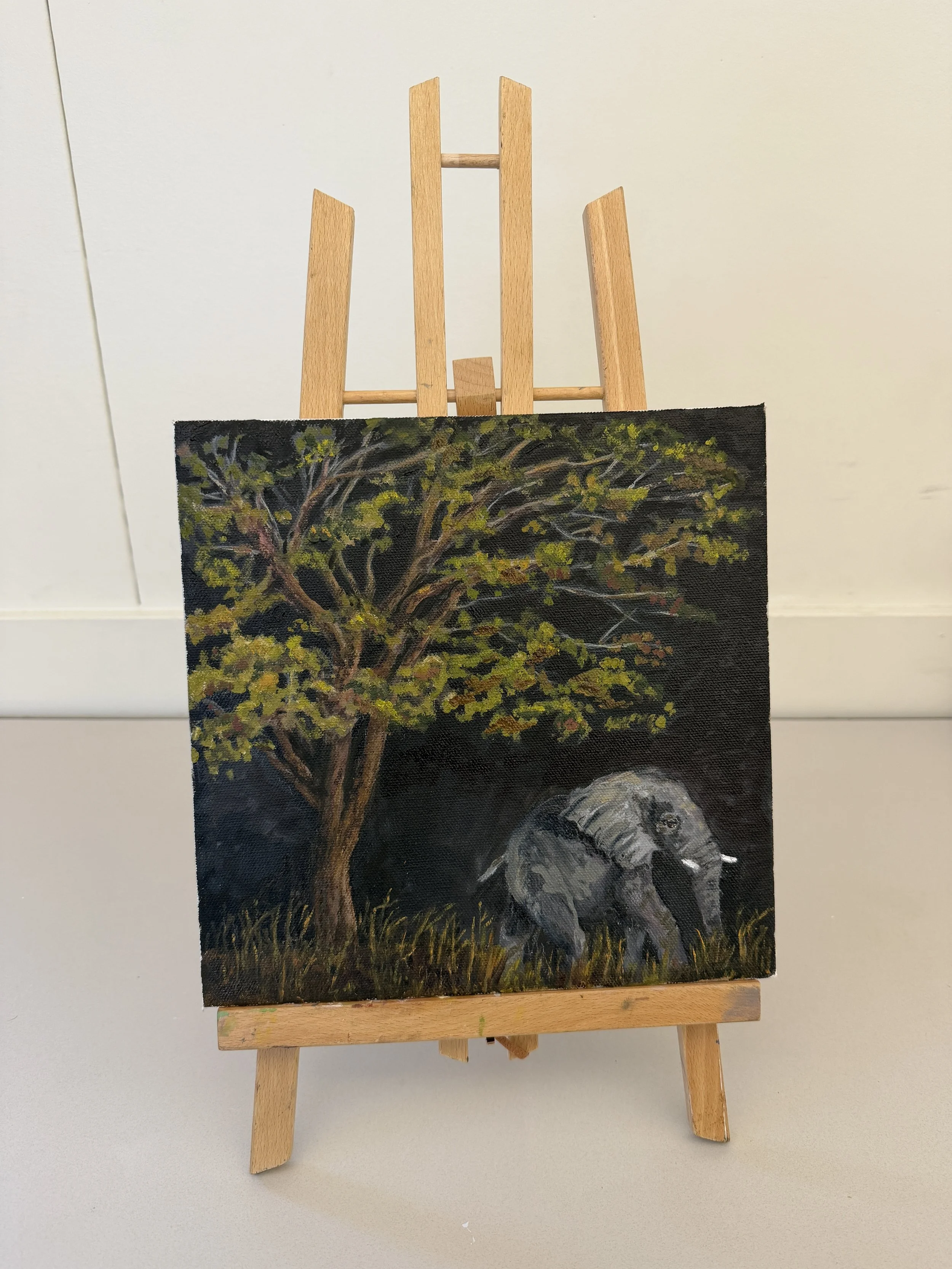 A small painting of an elephant standing under a large tree, with grass at its base, on an easel set against a plain wall.