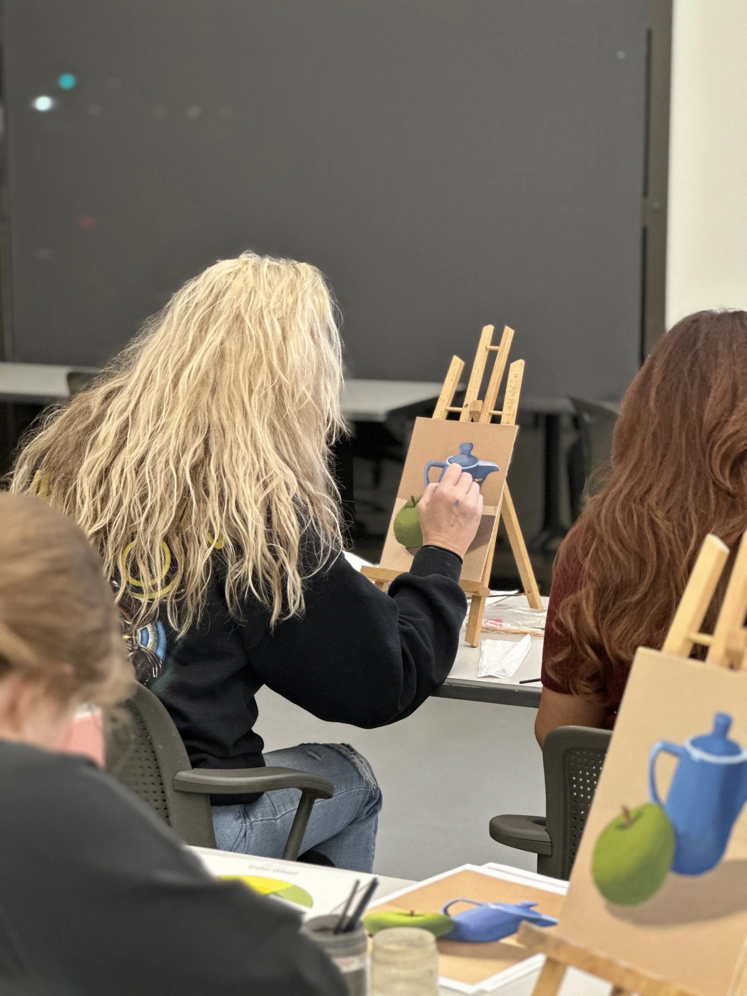A woman with curly blonde hair seated at a table painting on a canvas, with an easel showing a teapot and two pears, in a classroom or workshop setting with people seated nearby.