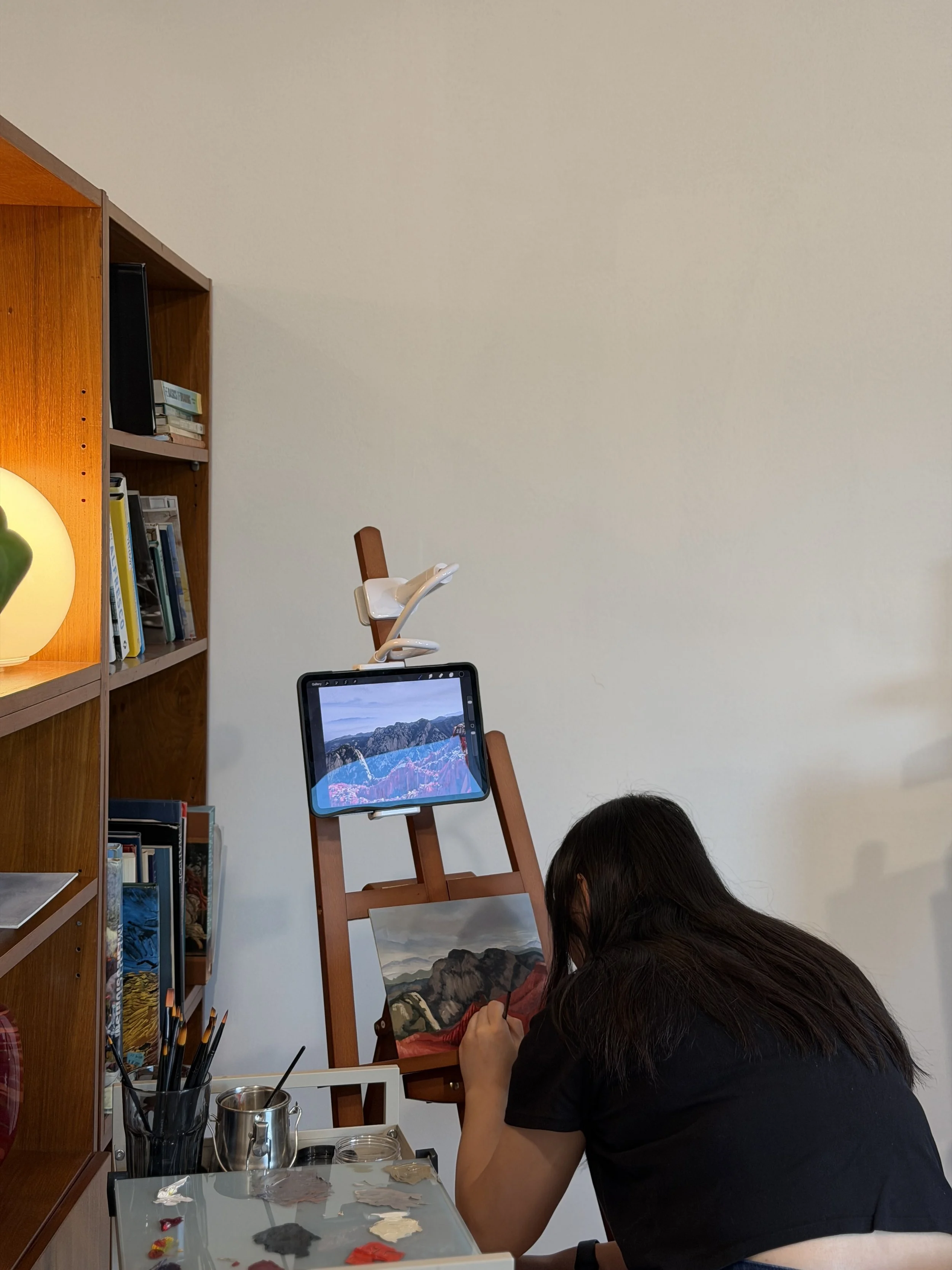 Person painting a landscape on a canvas set on an easel in a room with shelves and art supplies.