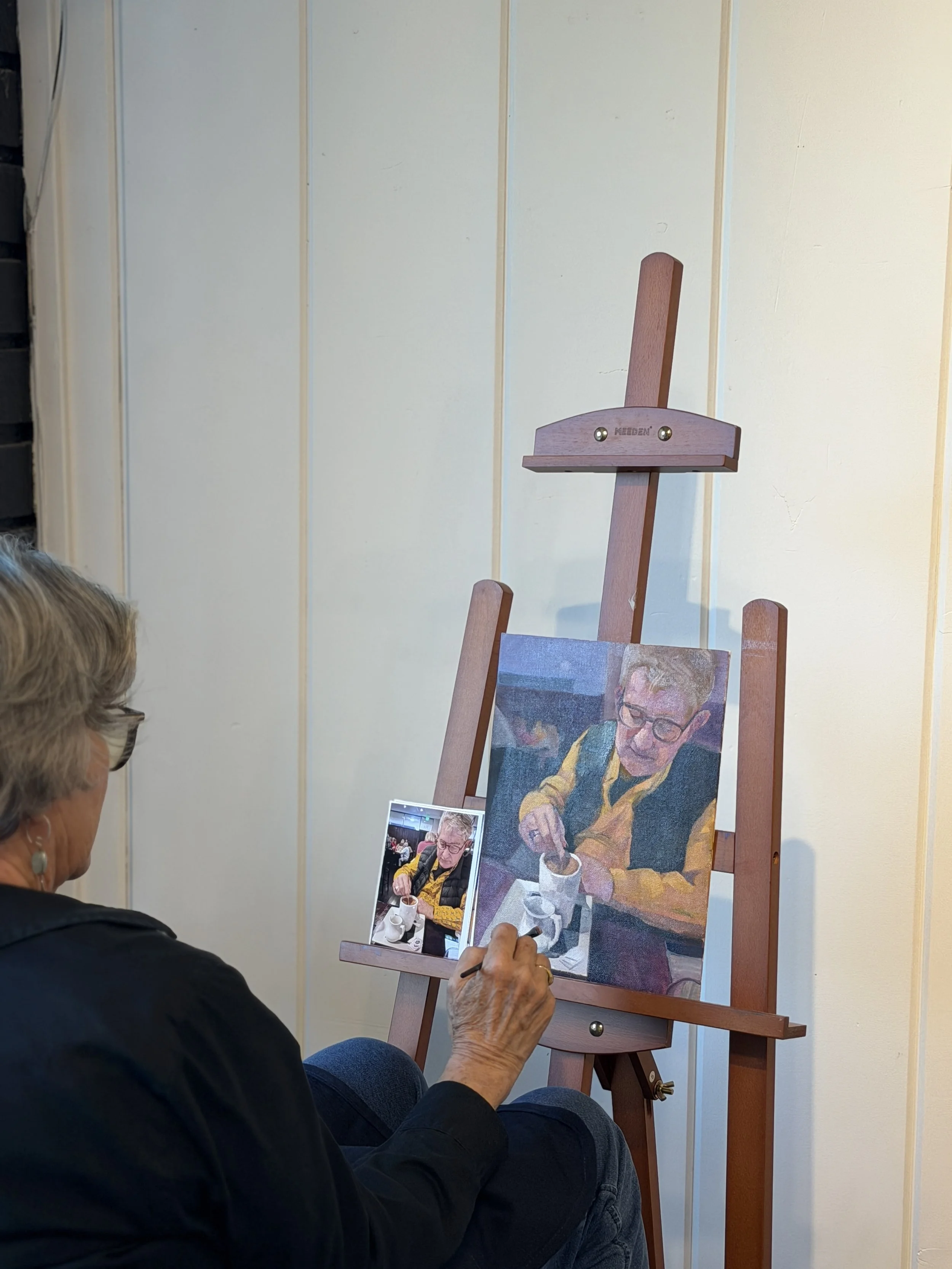 An older woman is seated, holding a small painted portrait easel displaying a portrait of a person with short blonde hair, glasses, and a yellow jacket, drinking from a cup.