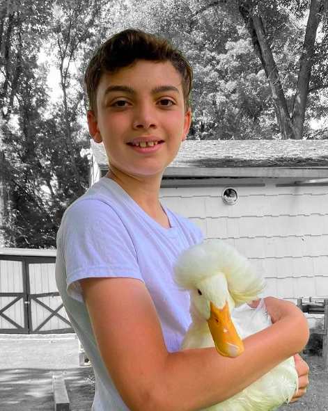 A young boy smiling and holding a white duck outdoors in front of a house and trees.