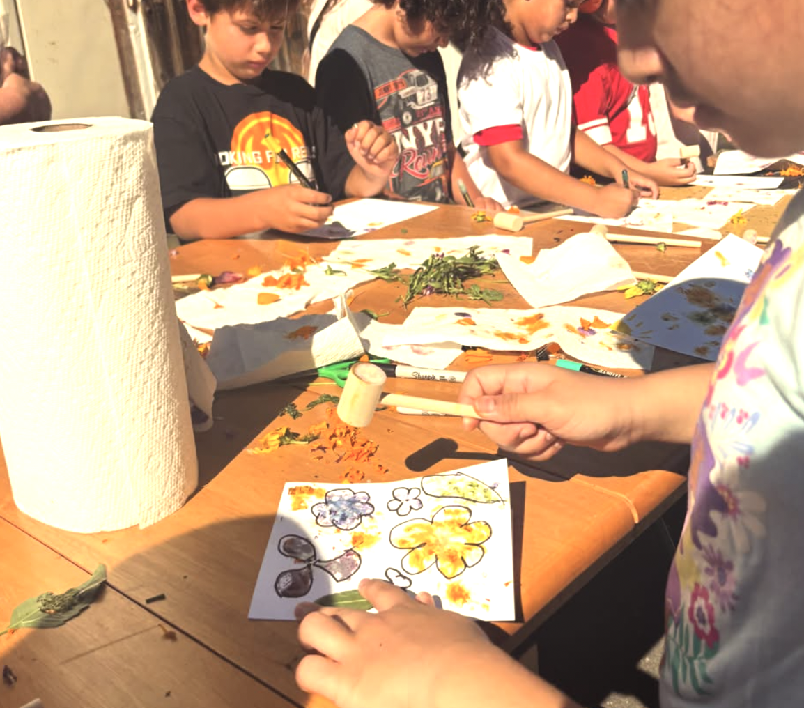 Children engaged in an outdoor arts and crafts activity at a wooden table, with various art supplies and colorful flower drawings on paper.