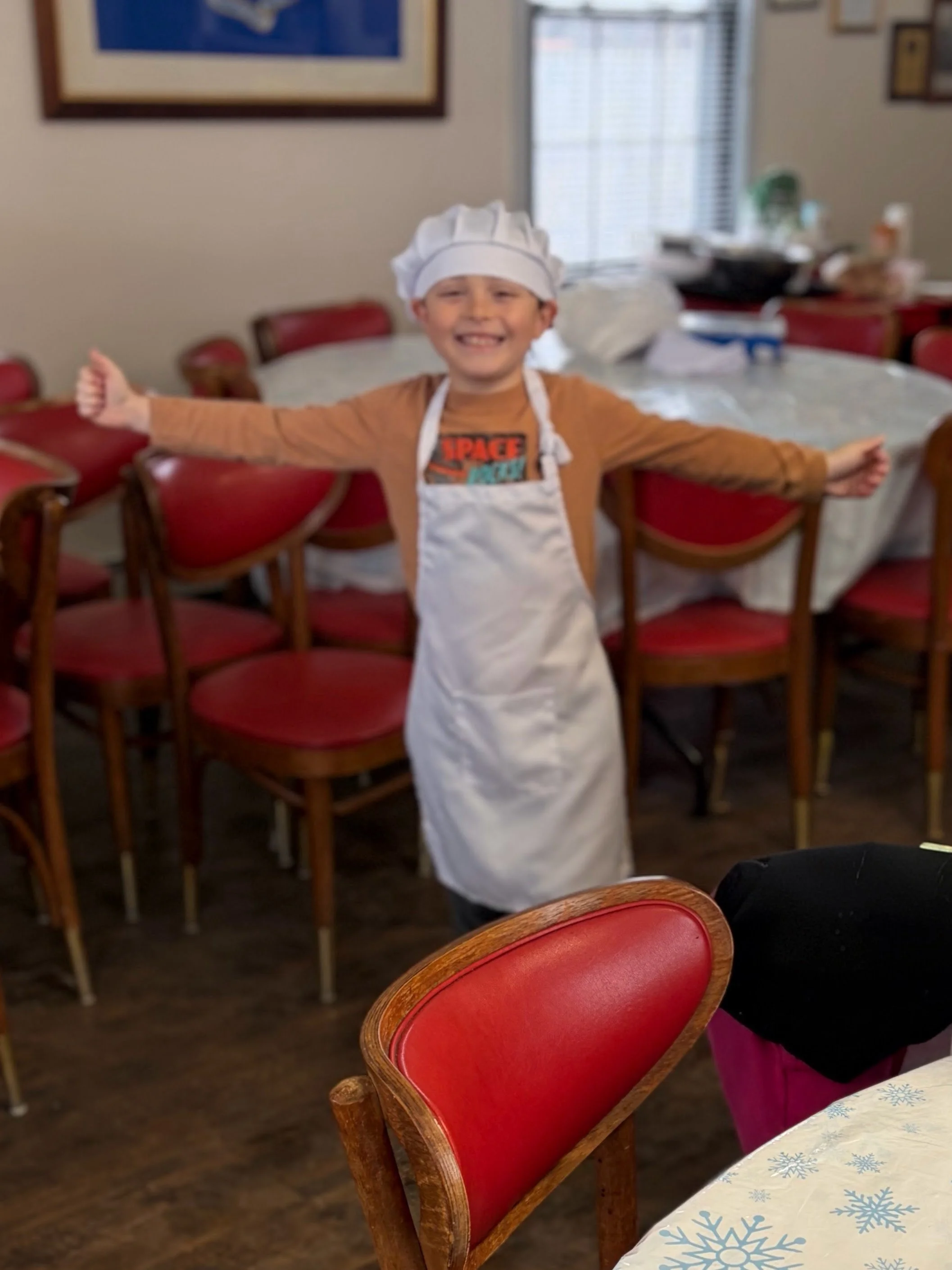 A young boy wearing a white chef's hat and apron stands in a dining room with red chairs and a table, smiling with arms outstretched.