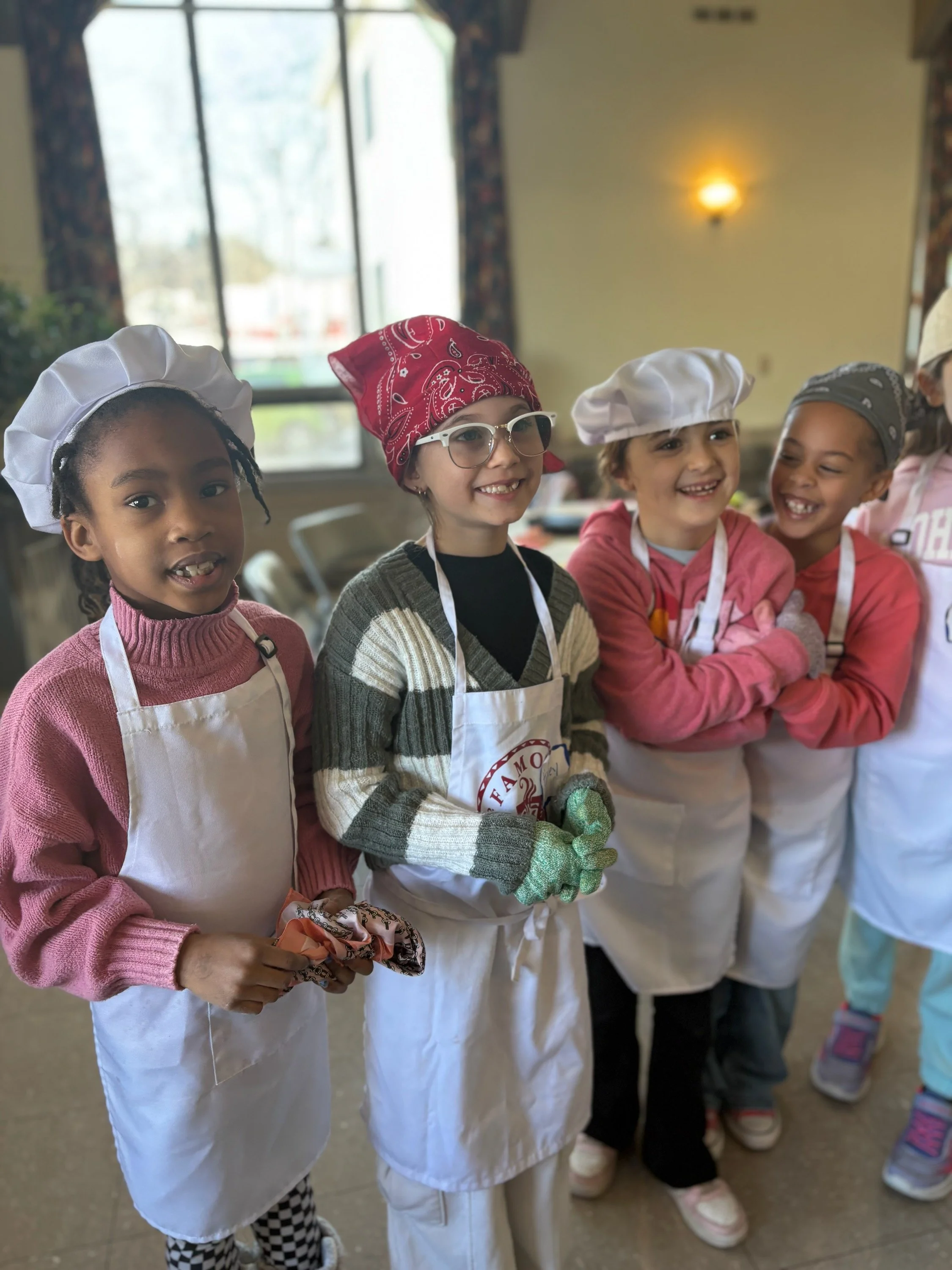 Group of children wearing chefs' hats and aprons, smiling in a kitchen or cooking class.