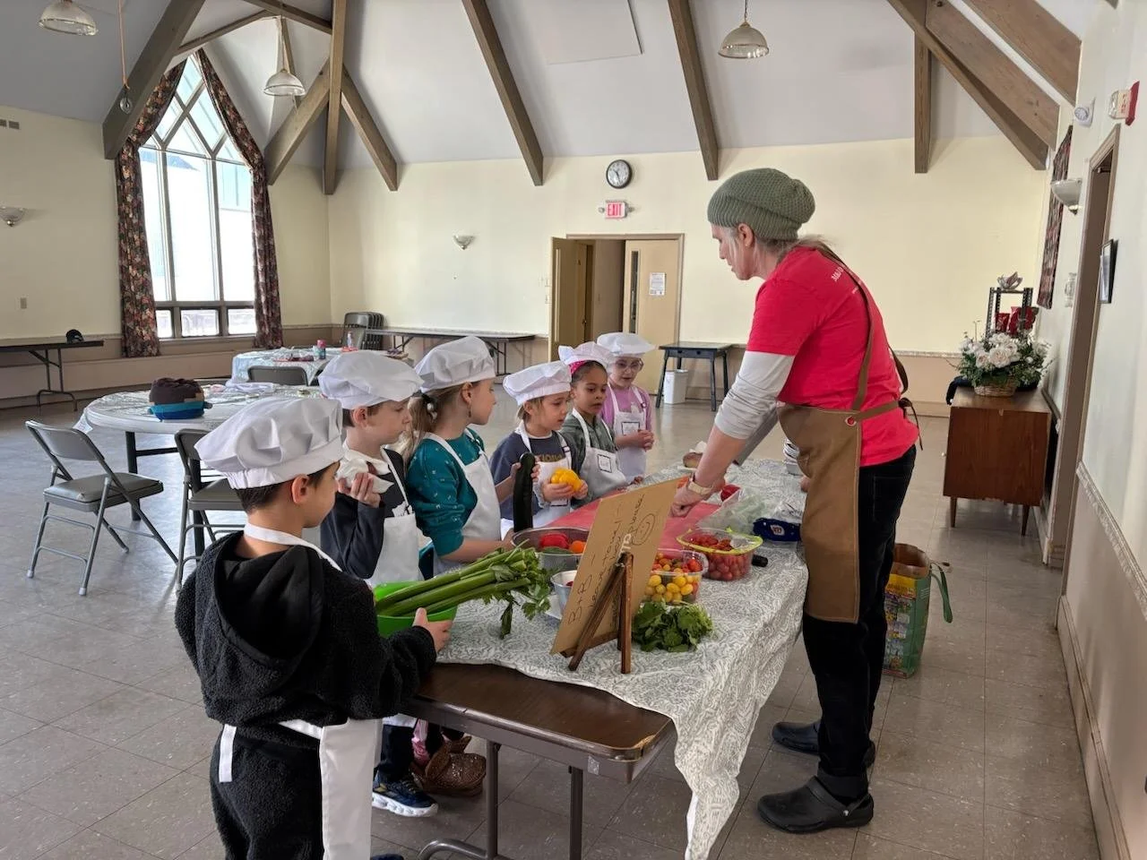 A woman demonstrating cooking or food preparation to a group of children dressed as chefs in a community hall.