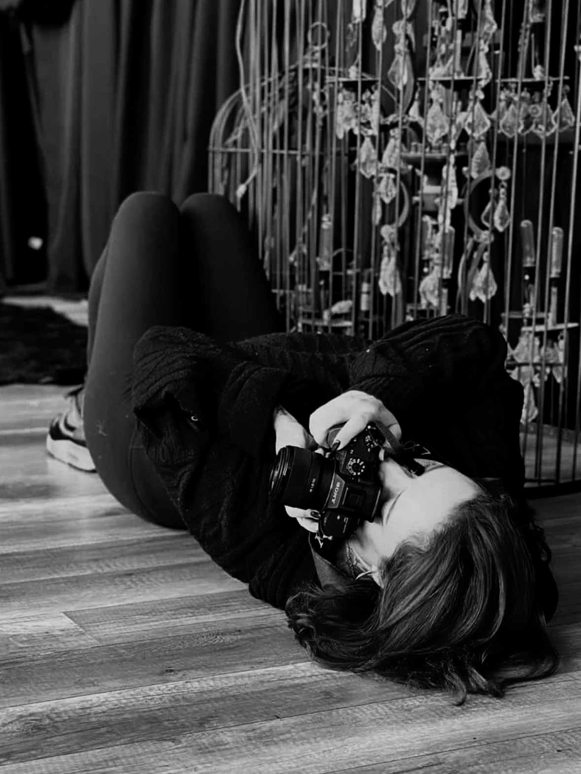 Person lying on the floor holding a camera with a floral cage in the background.