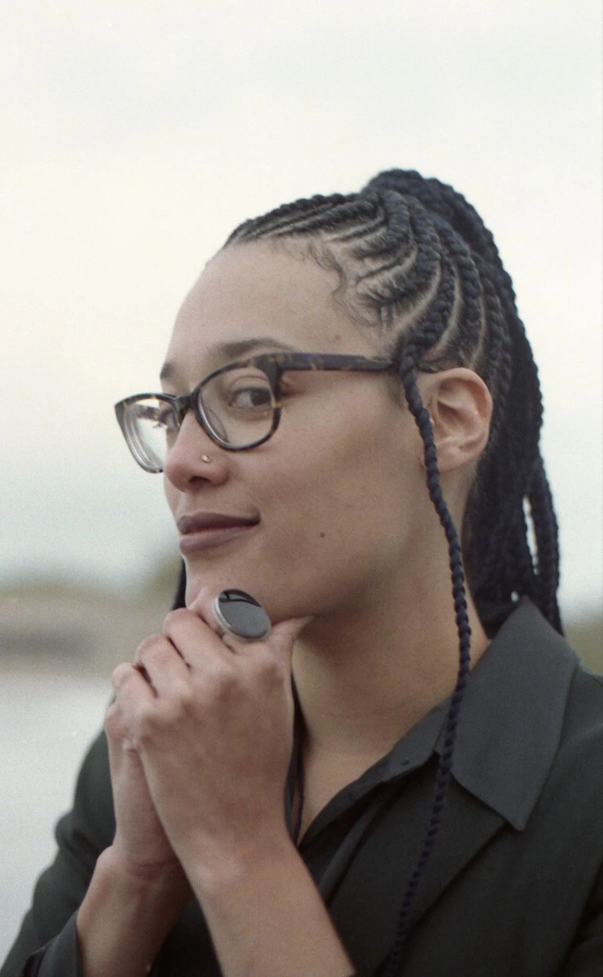Dr. Fushcia-Ann Hoover in brown tortoise shell glasses, cornrows, and visible nose ring. Her chin is resting on folded hands and a black oval ring is on the pointer finger. She is wearing a black botton-down blouse.