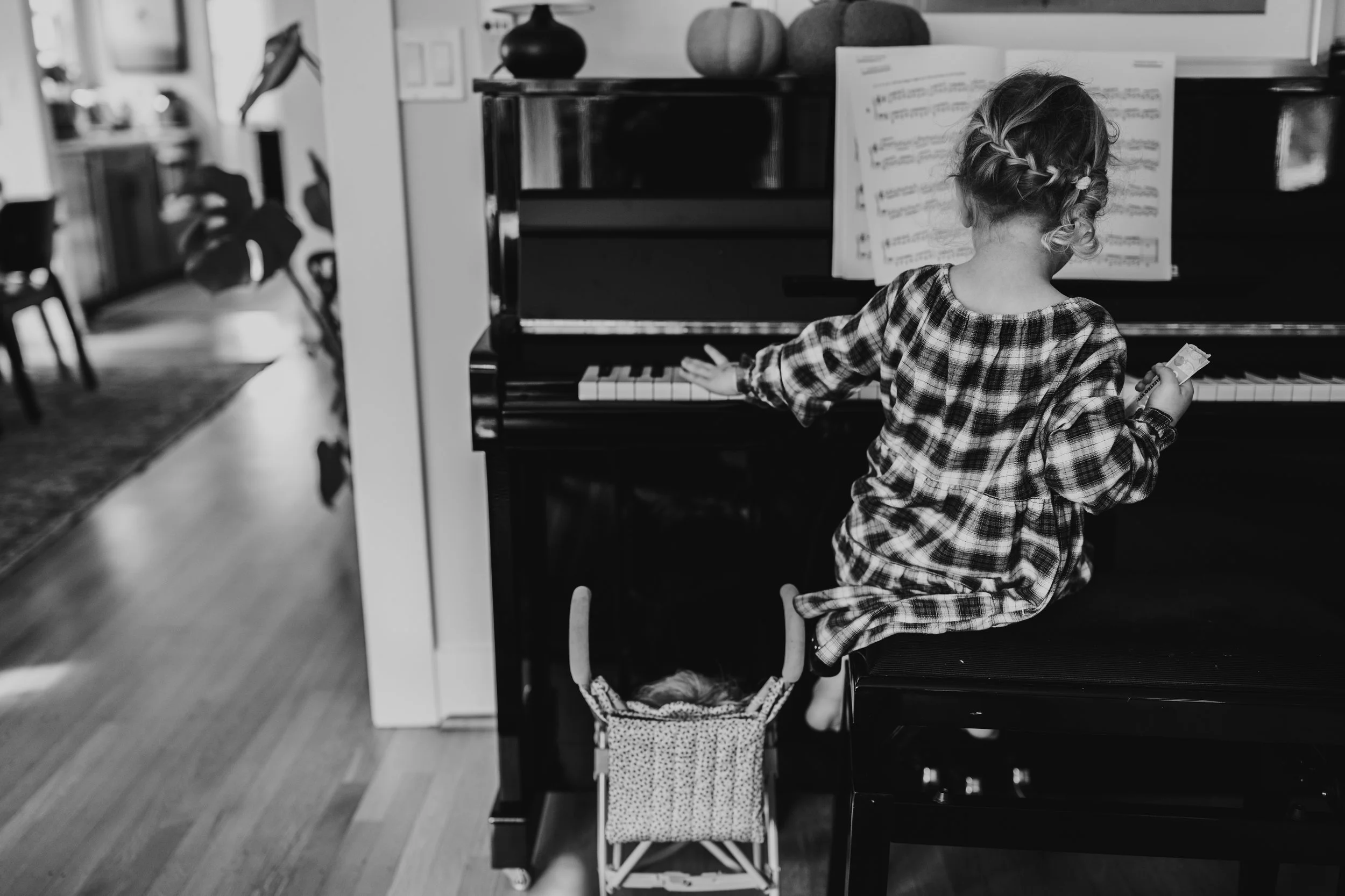 A young girl with braided hair playing the piano in a home setting, wearing a checkered dress, and holding a small object in her hand.
