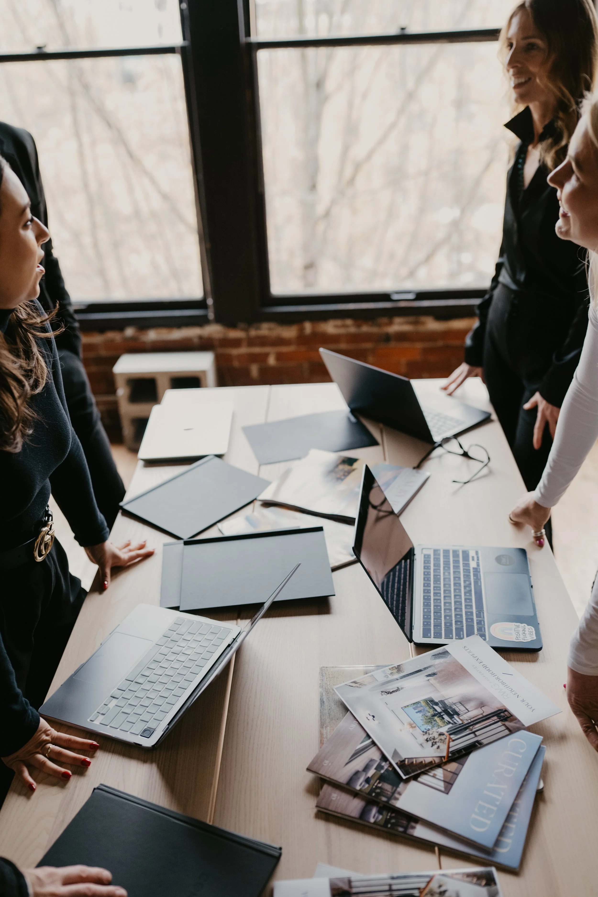 A group of women gathered around a table with laptops, notebooks, and magazines during a meeting or discussion in a bright room with large windows.